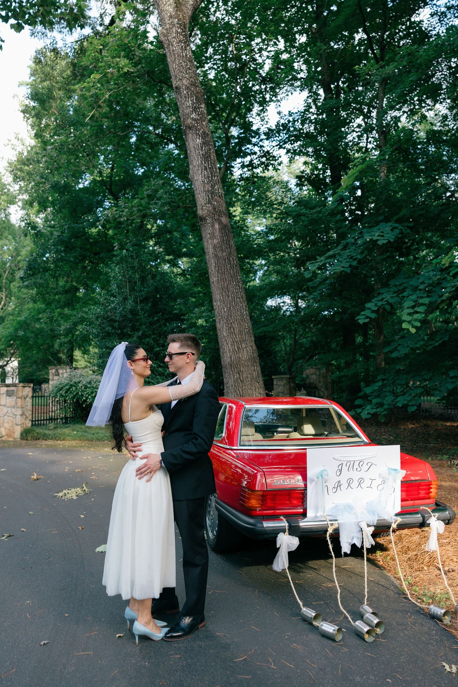 Bride in a short white dress and veil embraces her groom beside a red vintage car with a Just Married sign and tin cans.