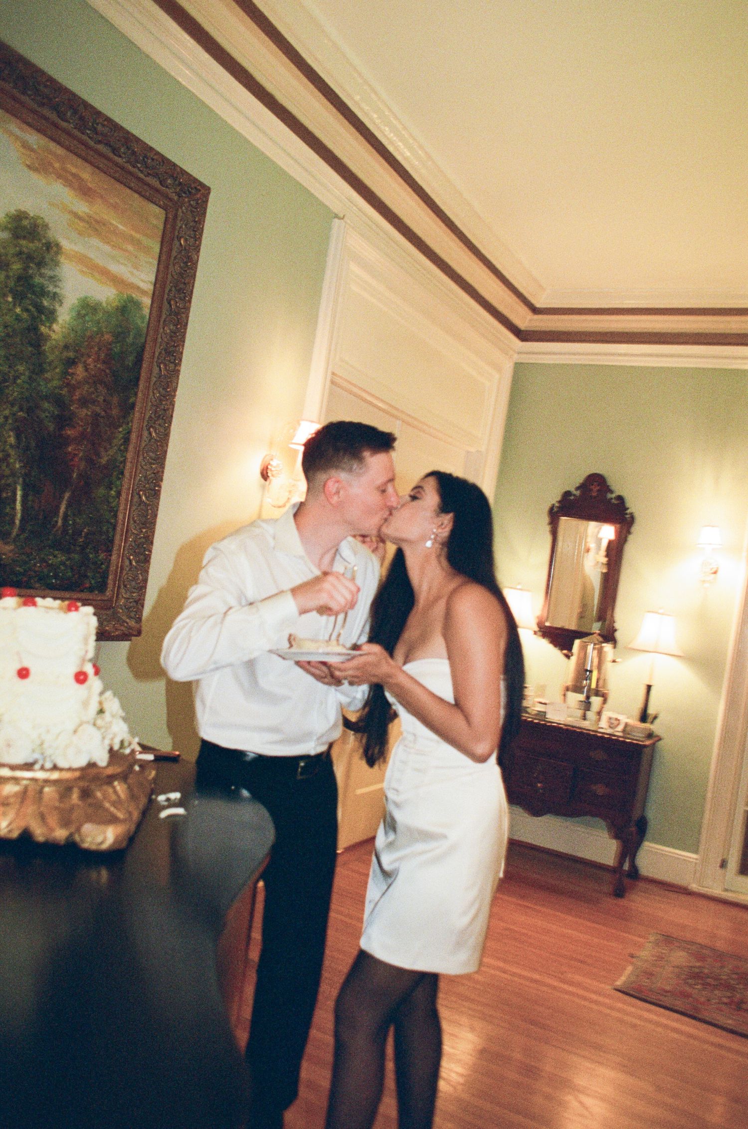 Couple in white kissing while holding a plate of wedding cake beside a tiered cake in an elegant room with wood floors.