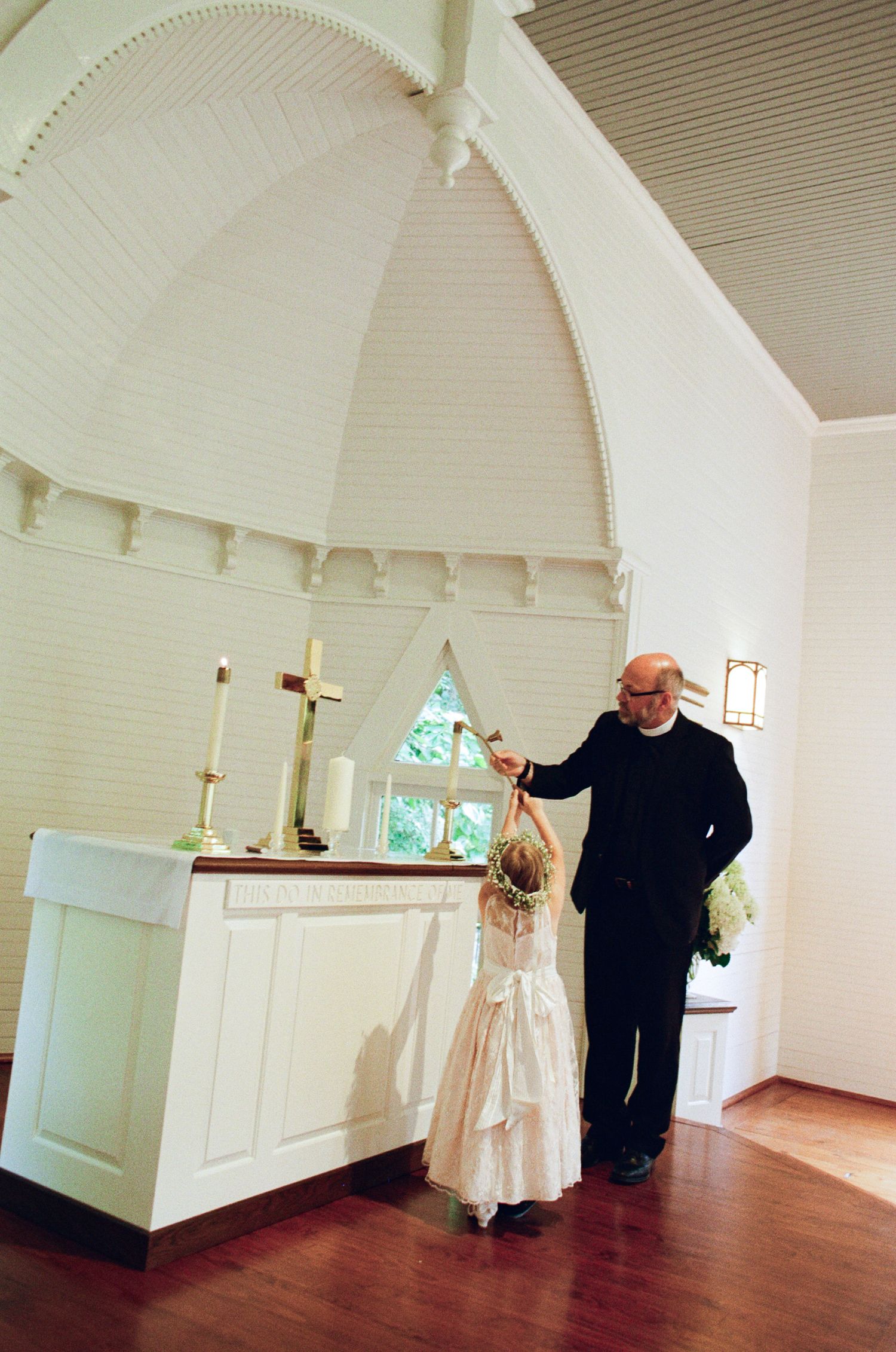 A flower girl in a white dress reaches toward a candle on the altar as a minister in black watches in a bright white chapel.
