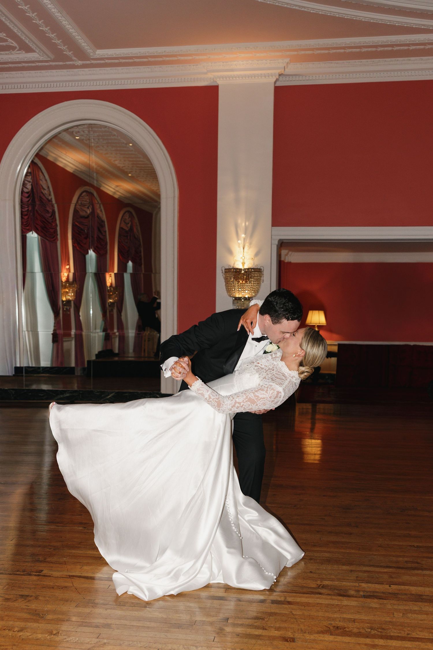 Bride and groom share a romantic dip kiss on the dance floor of an elegant red ballroom during their first wedding dance.