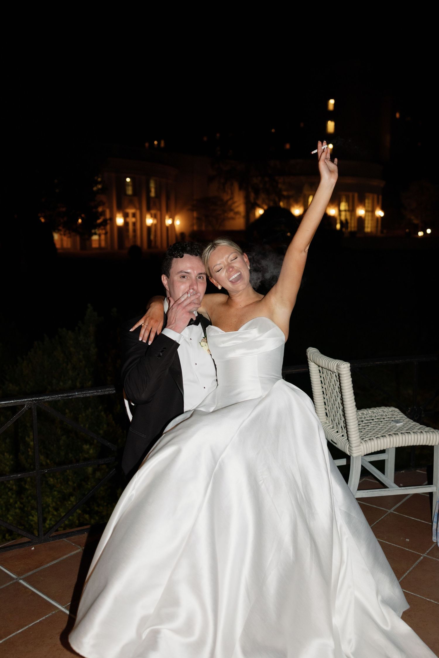 Joyful bride in white gown raises arm while groom in tuxedo poses beside her at night wedding celebration.