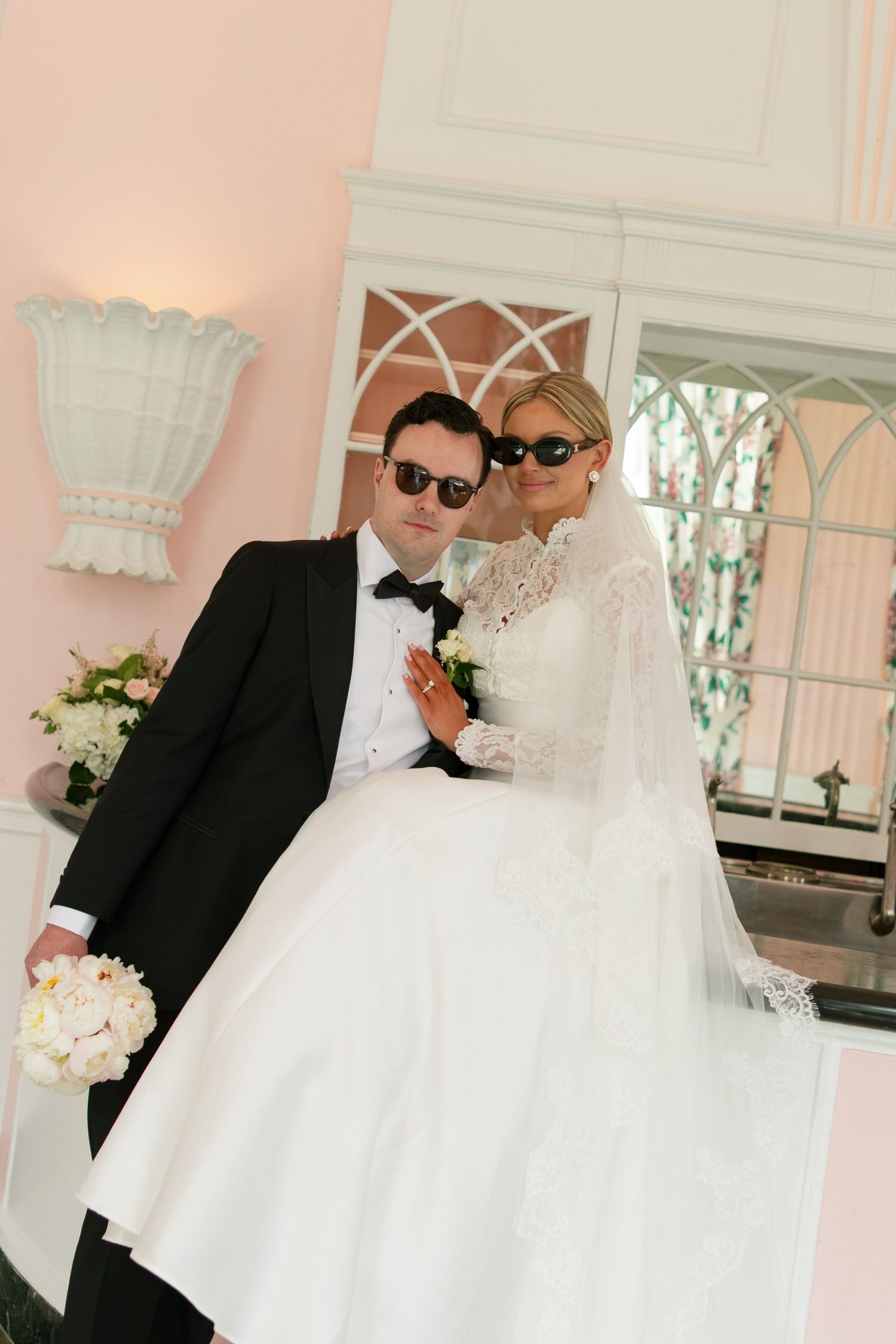 Elegant bride and groom in sunglasses posing together outside a pink building on their wedding day.