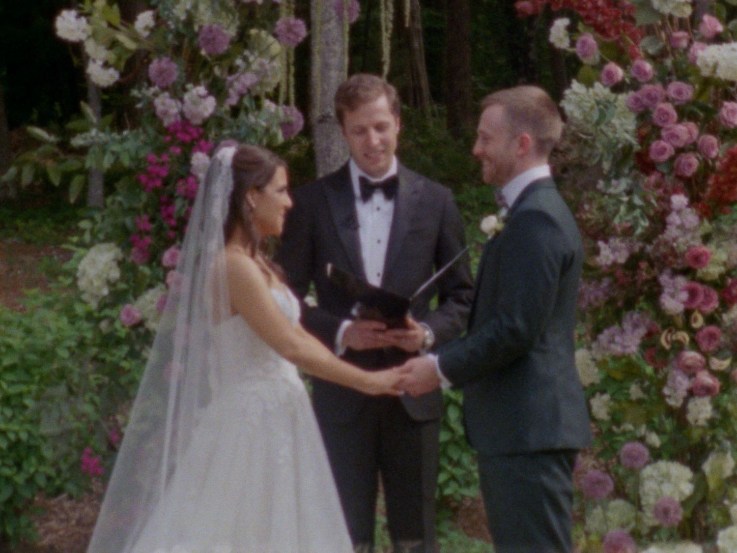 Bride and groom exchange vows outdoors at floral arch ceremony with officiant holding black book.