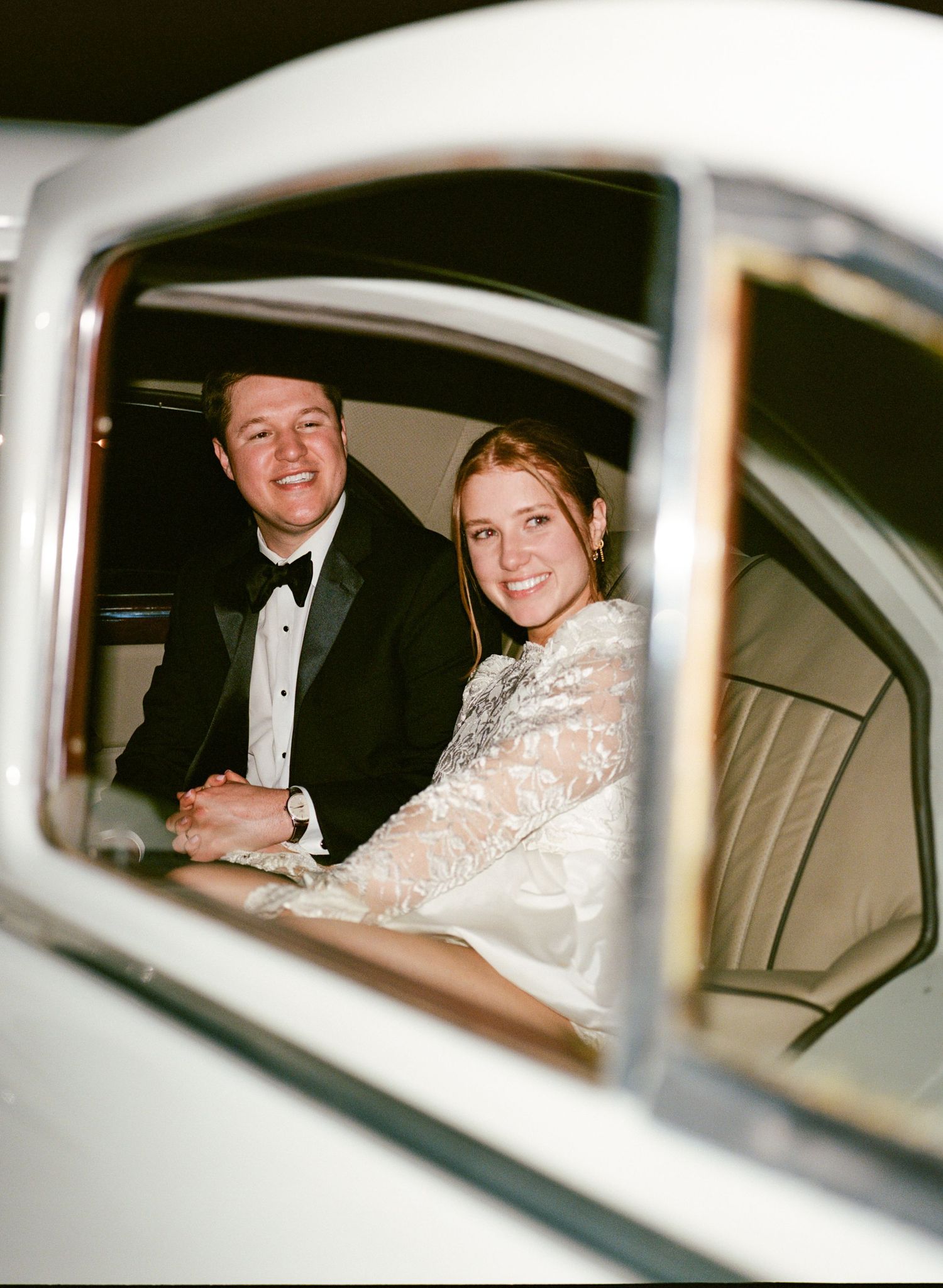 Bride and groom smiling together inside a white wedding car, man in tuxedo, woman in lace bridal gown.