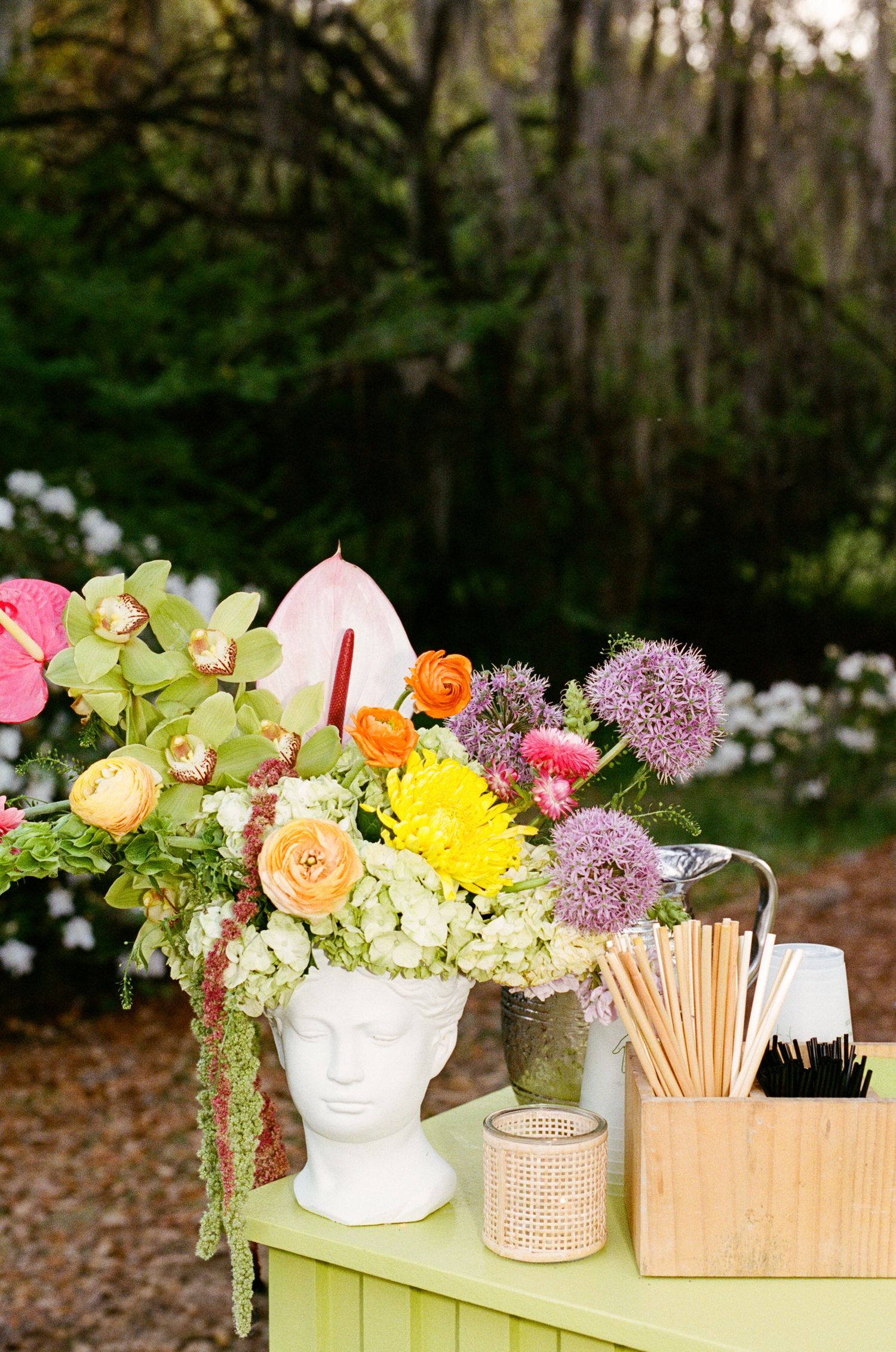 Colorful floral arrangement in white head vase on green table with wooden box of straws outdoors.