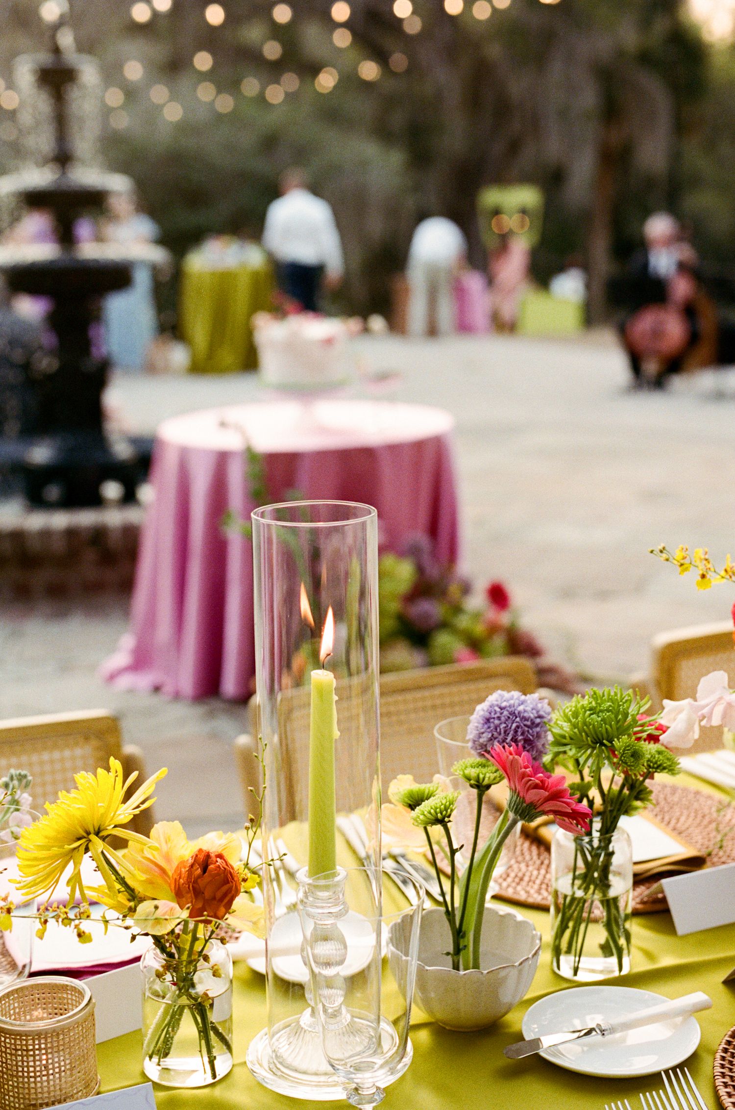 Elegant outdoor wedding reception table with green candles, colorful flowers, and pink tablecloths in a garden setting.