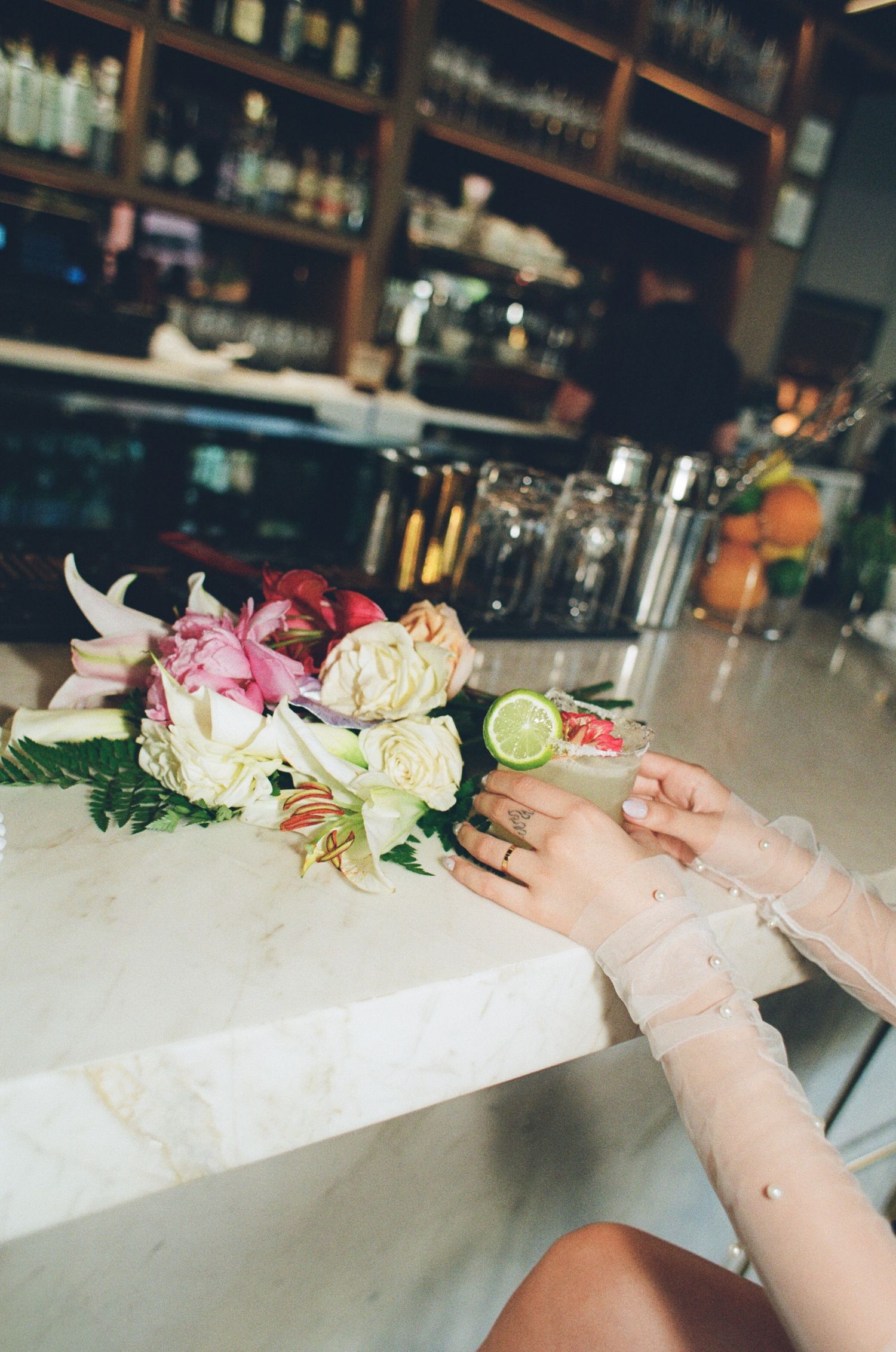 Hands wearing gloves arrange colorful tropical flowers and garnishes on a bar counter filled with crushed ice.