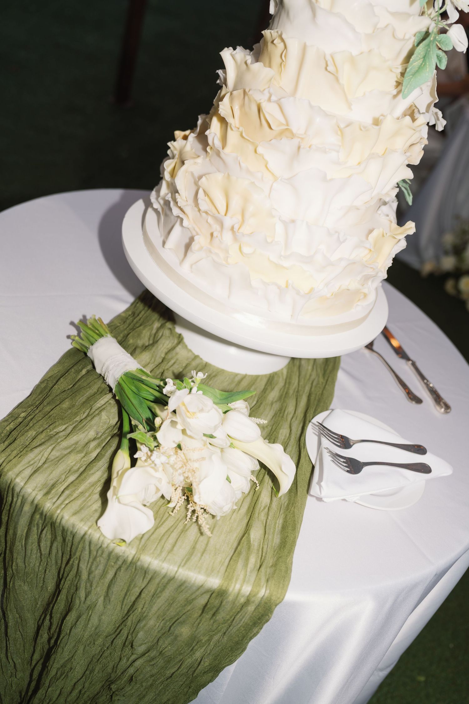 A white wedding cake sits on a table with green fabric runner and white floral decorations.