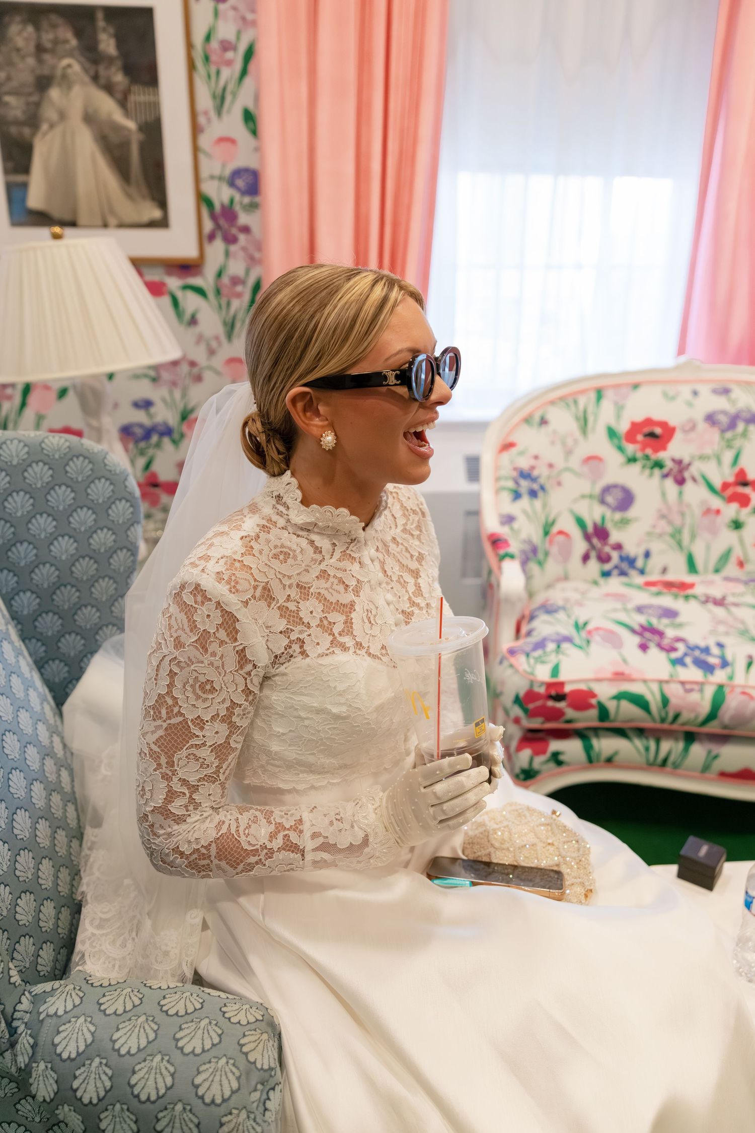 Bride in white lace gown and sunglasses sitting in floral chair, holding cake slice and drink at wedding reception.