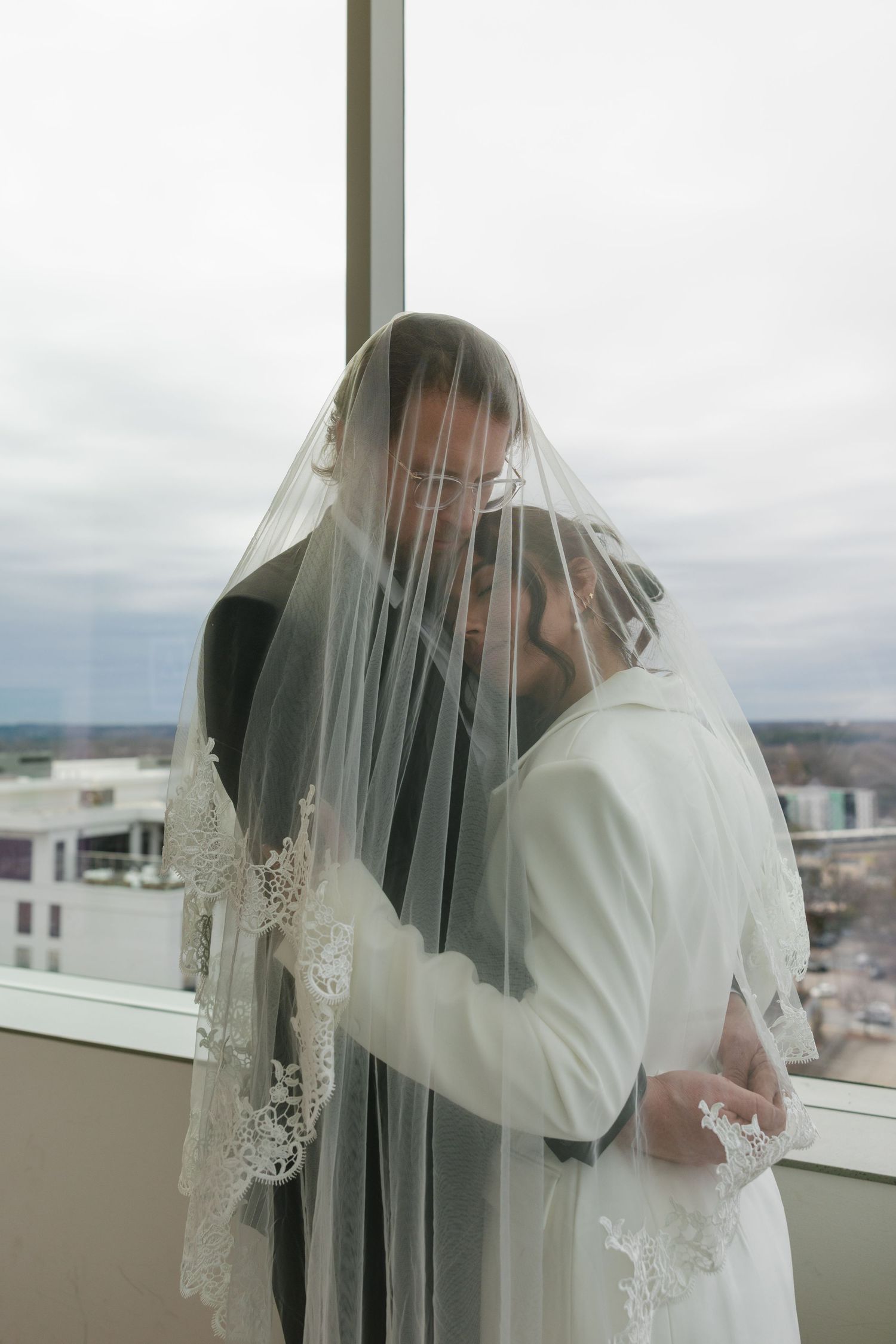 Bride and groom share intimate moment under lace wedding veil by large window on their wedding day.