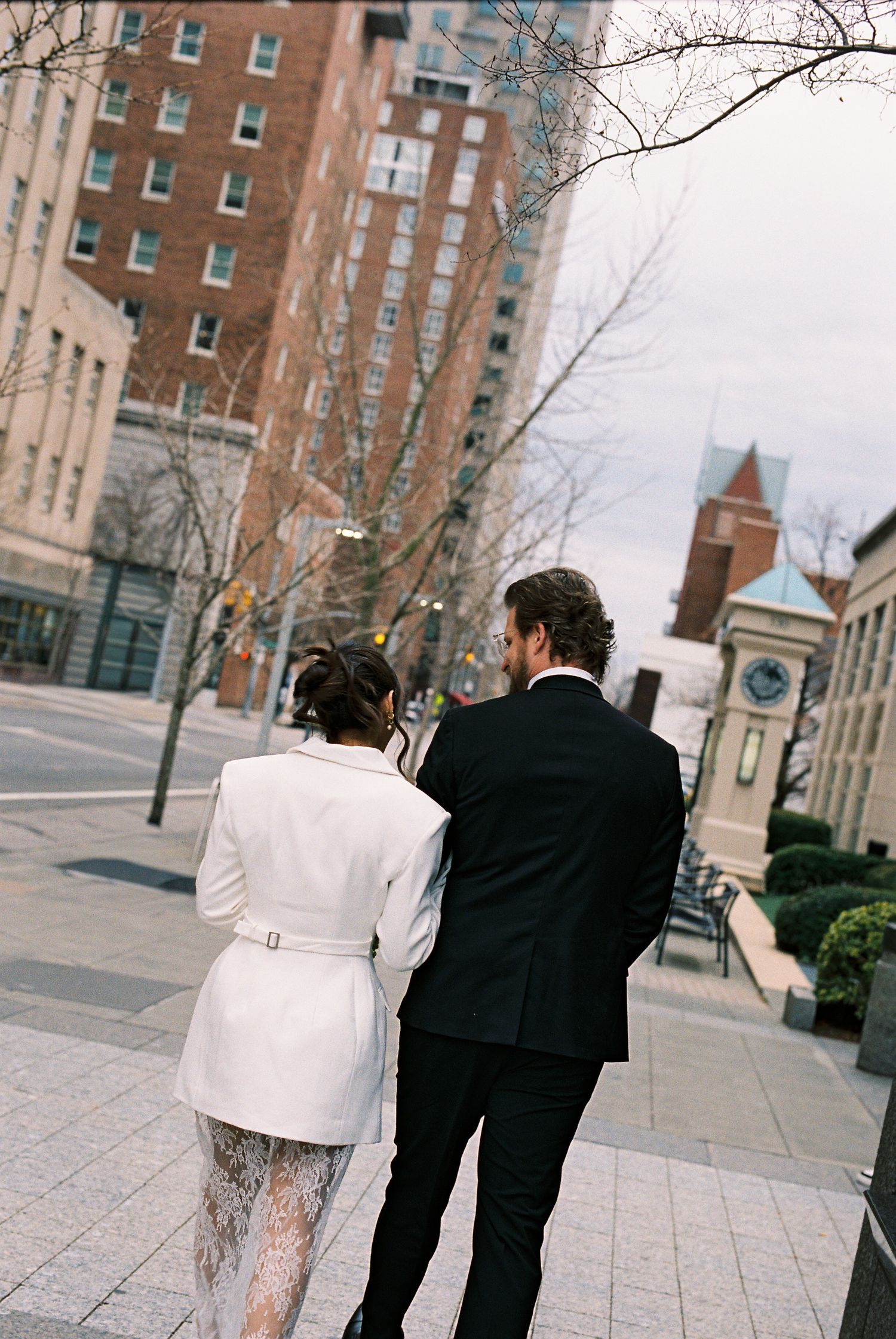 Couple walking through urban city street, woman in white coat and lace dress, man in black suit, tall buildings backdrop.