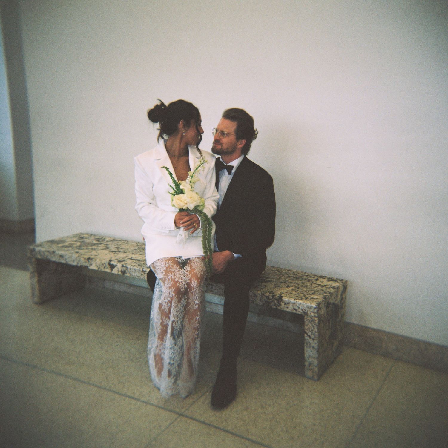 Interracial couple in wedding attire sitting on stone bench, bride holding white bouquet, groom in black suit and tie.