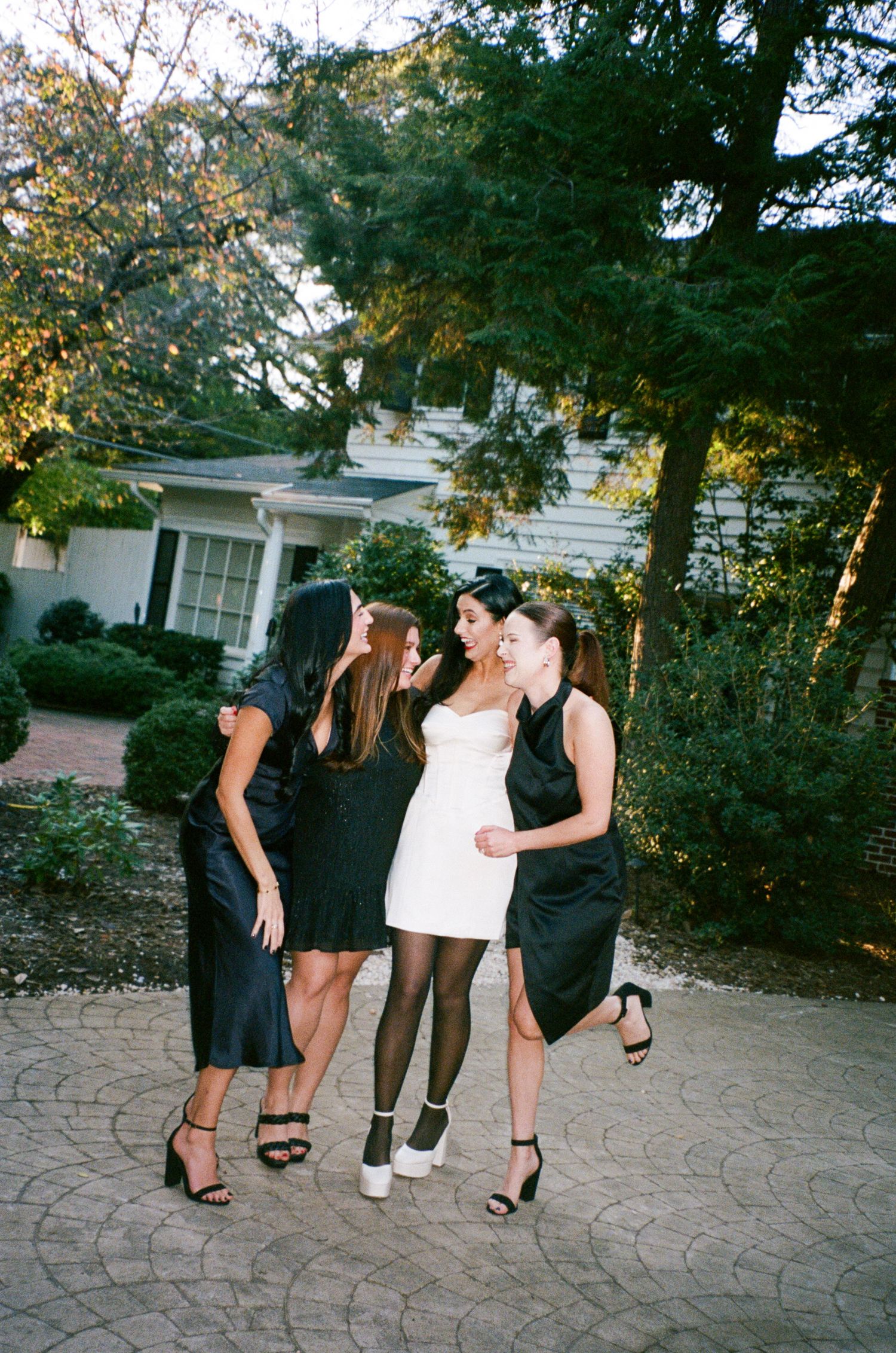 Four women in black and white cocktail dresses and heels laugh and huddle together on a brick patio in front of a house surrounded by trees.