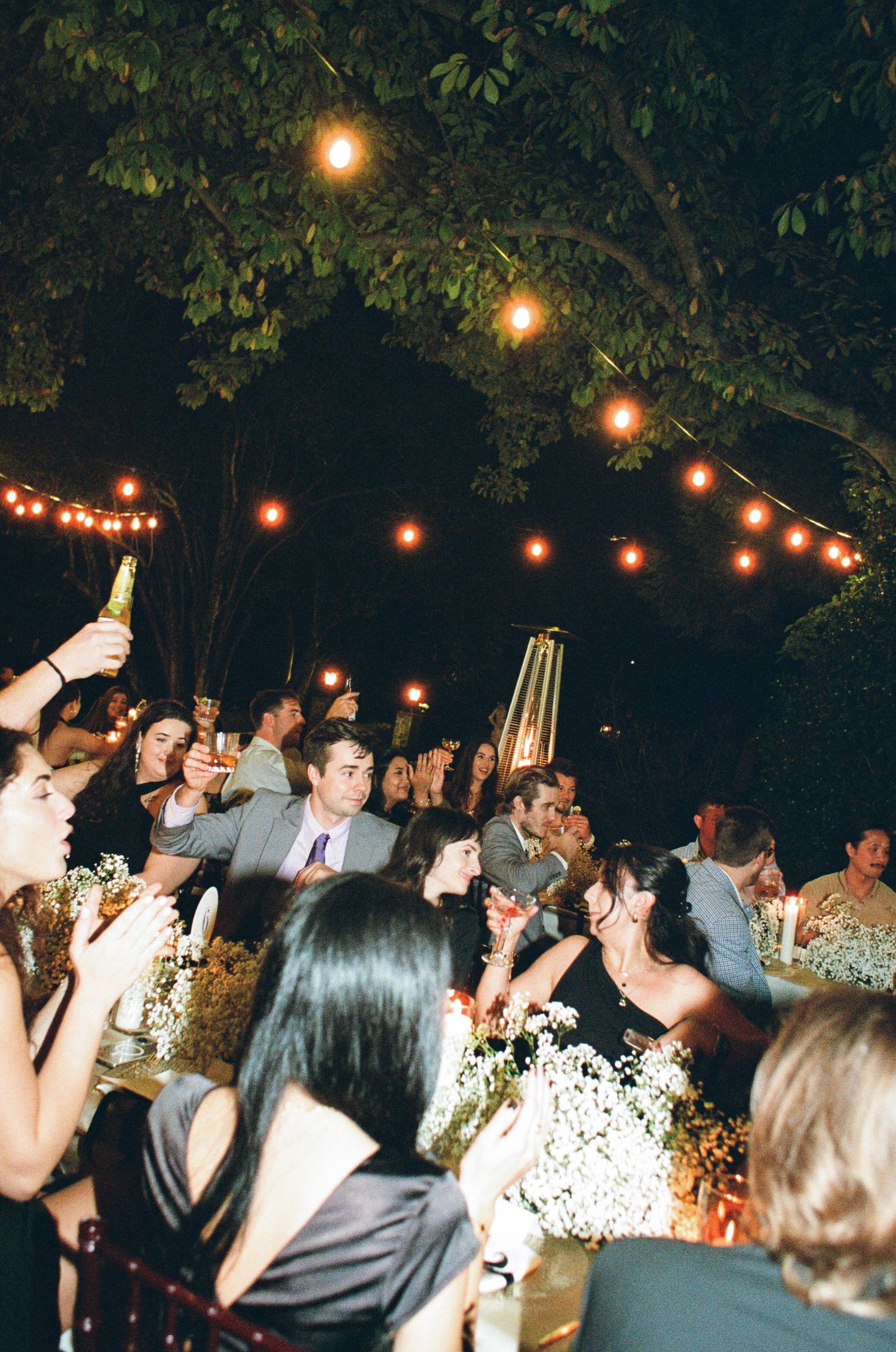 Wedding guests raise glasses in a toast at an outdoor evening reception under trees with warm string lights and white floral centerpieces.