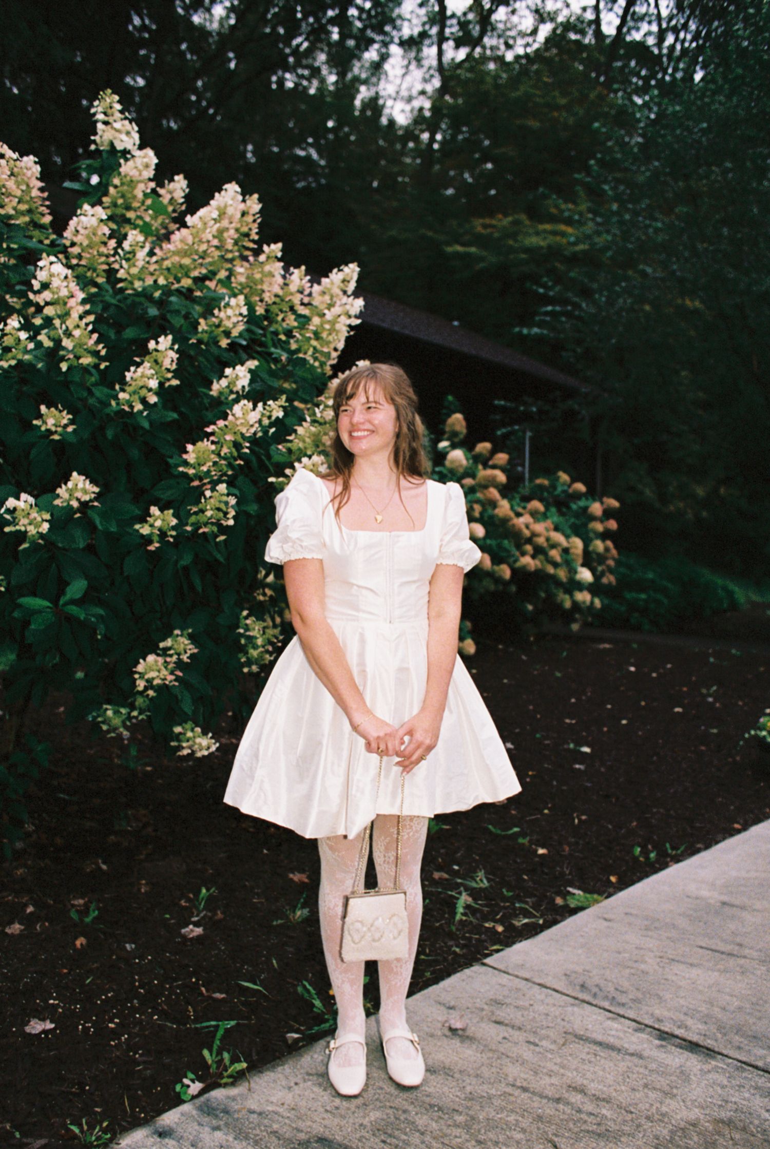 A person in a white vintage-style dress with puff sleeves stands by blooming hydrangea bushes on a garden path.
