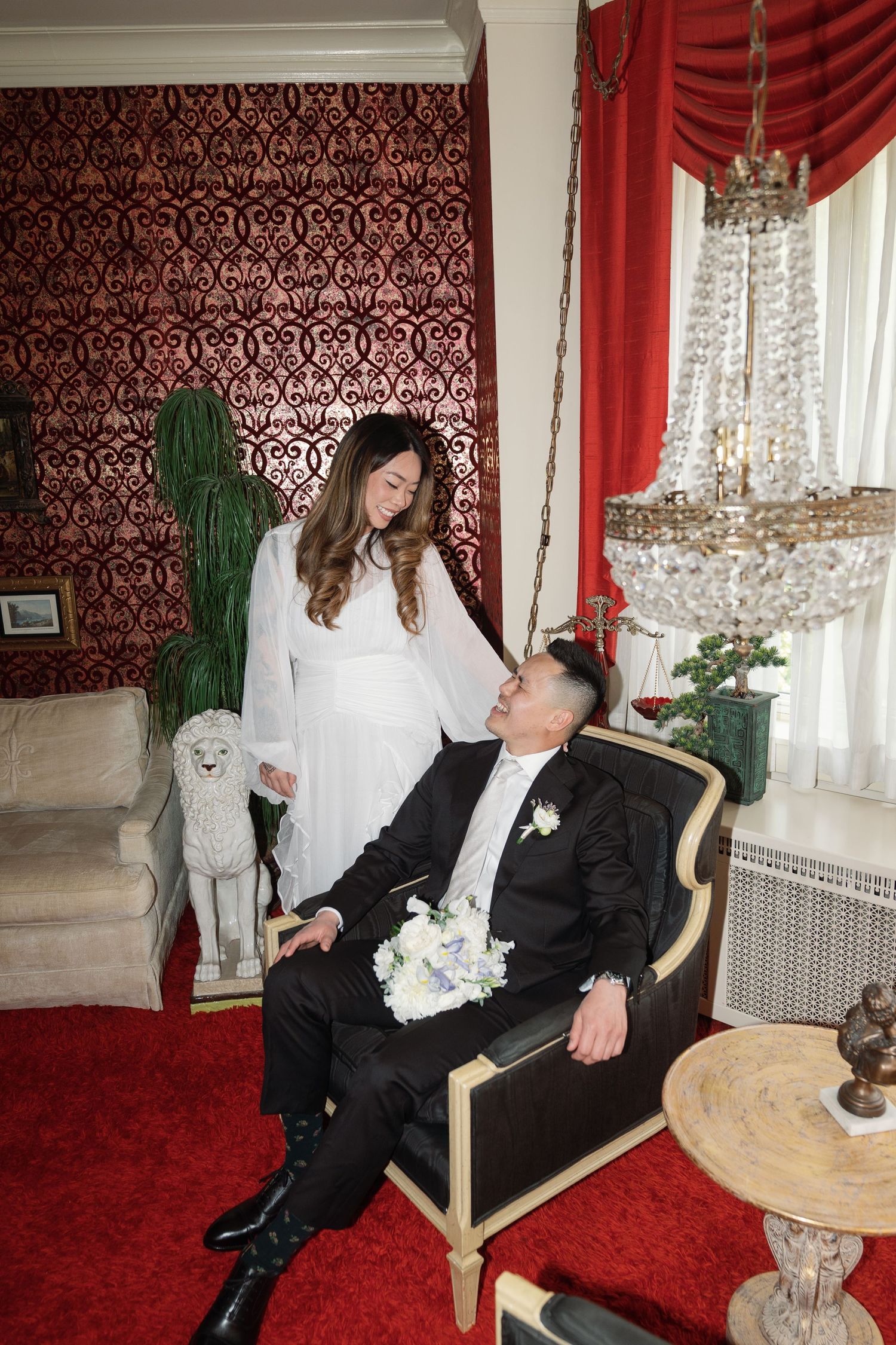 Elegant couple poses in luxurious room with red damask wallpaper, crystal chandelier and ornate furniture.