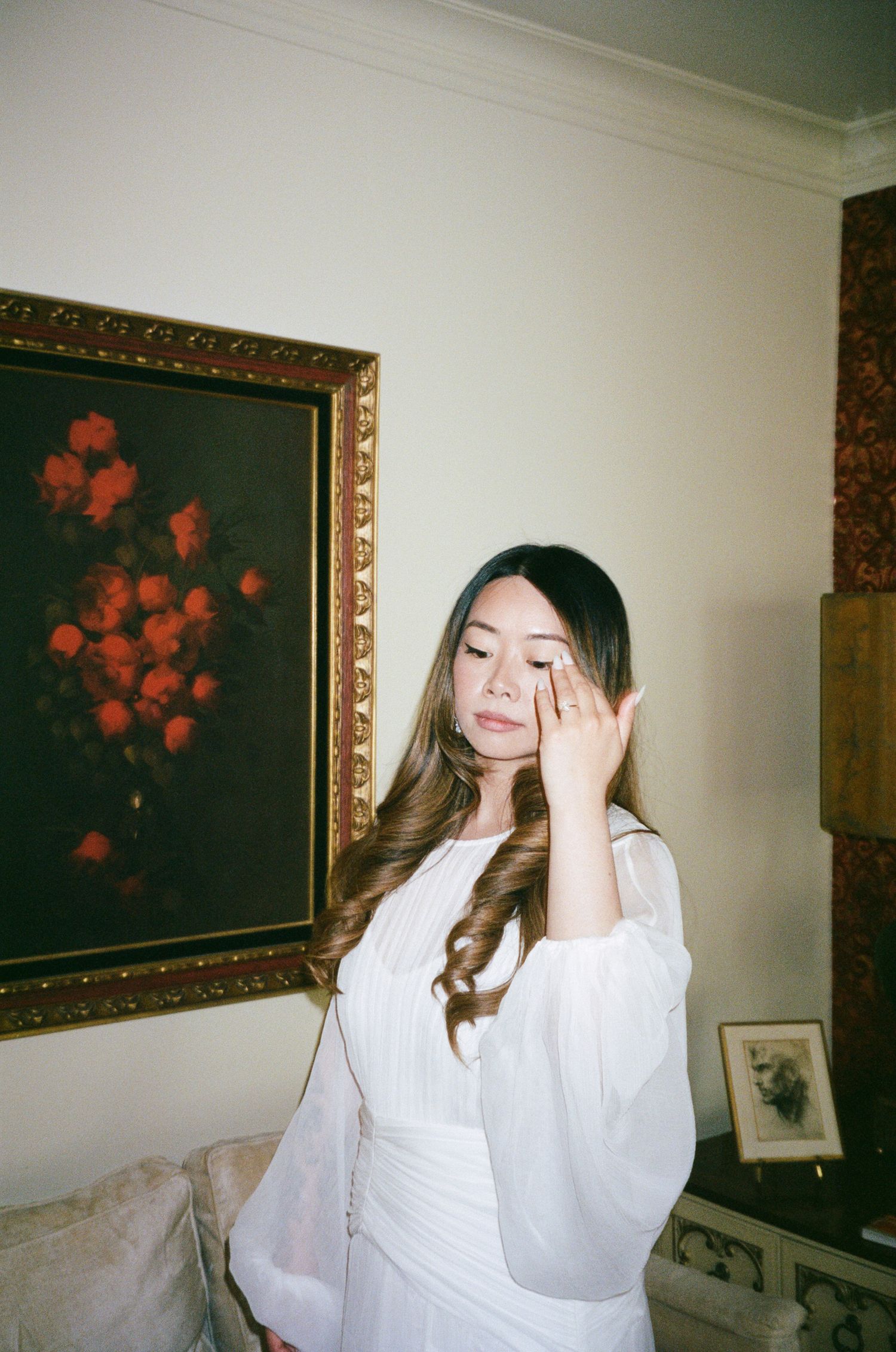 A person in a white flowing dress stands against a wall with a framed floral artwork in an ornate gold frame.
