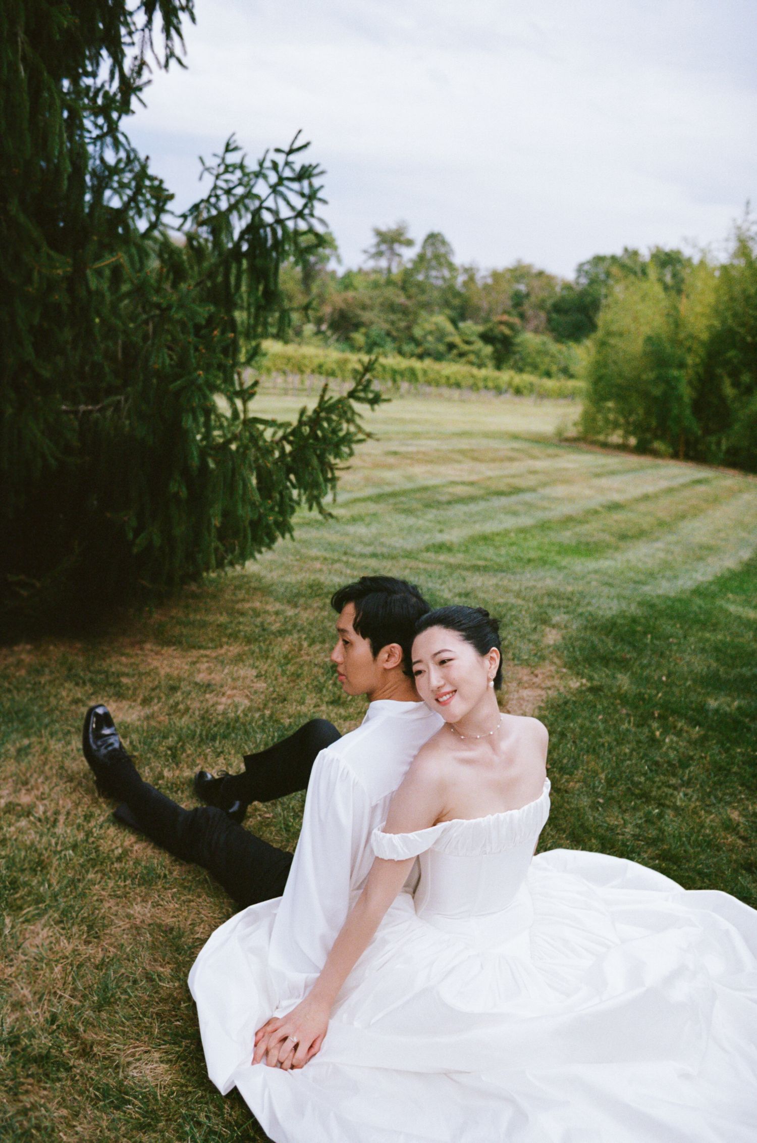 A couple in wedding attire sits together on grass taking a selfie in a scenic outdoor garden setting.