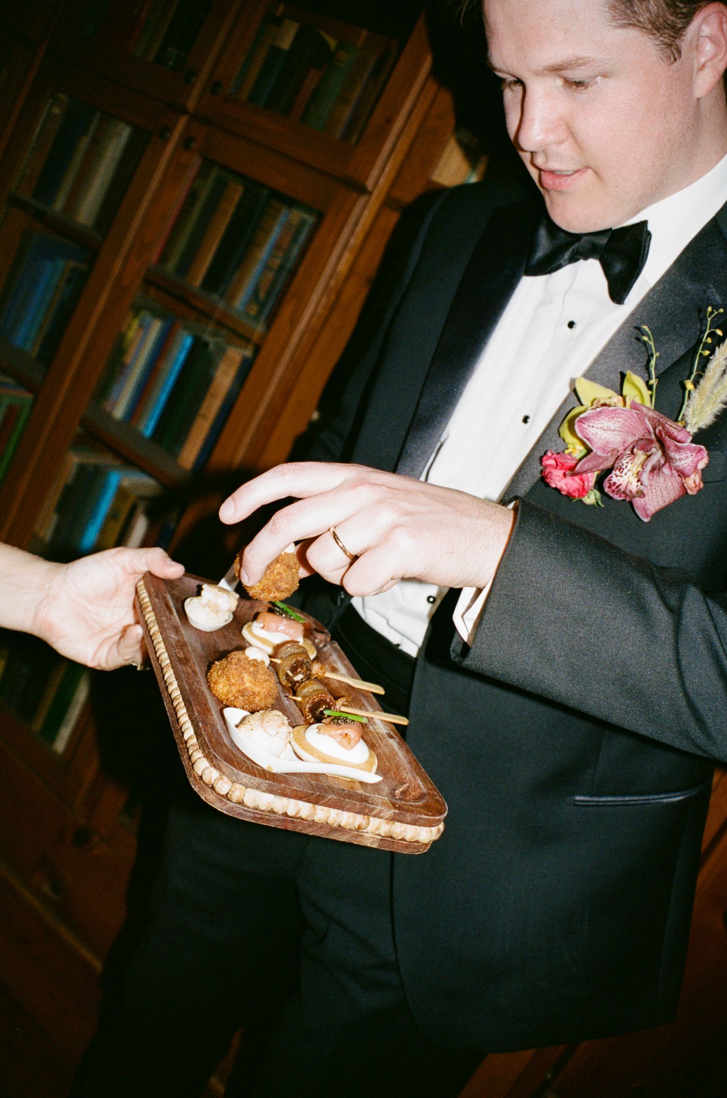 Groom in tuxedo with floral boutonniere selecting appetizers from a rustic wooden serving board at a wedding reception.