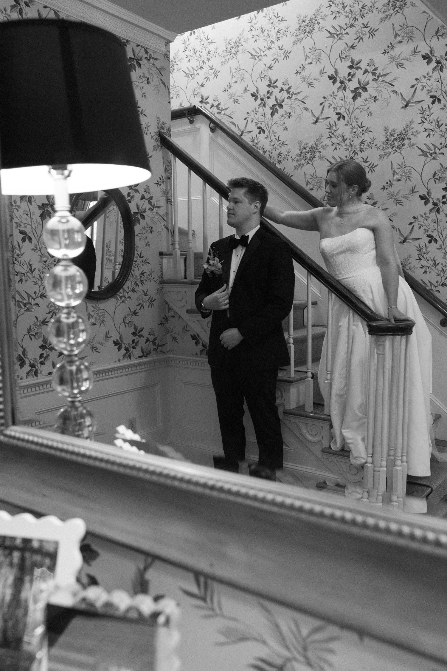 Black and white photo of bride and groom on elegant staircase with floral wallpaper during wedding day.