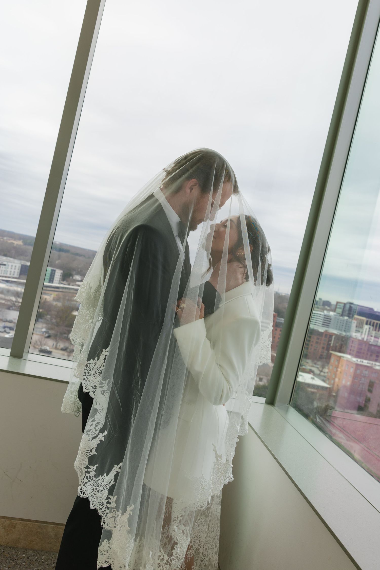 Bride and groom share intimate moment under lace veil by floor-to-ceiling windows on their wedding day.