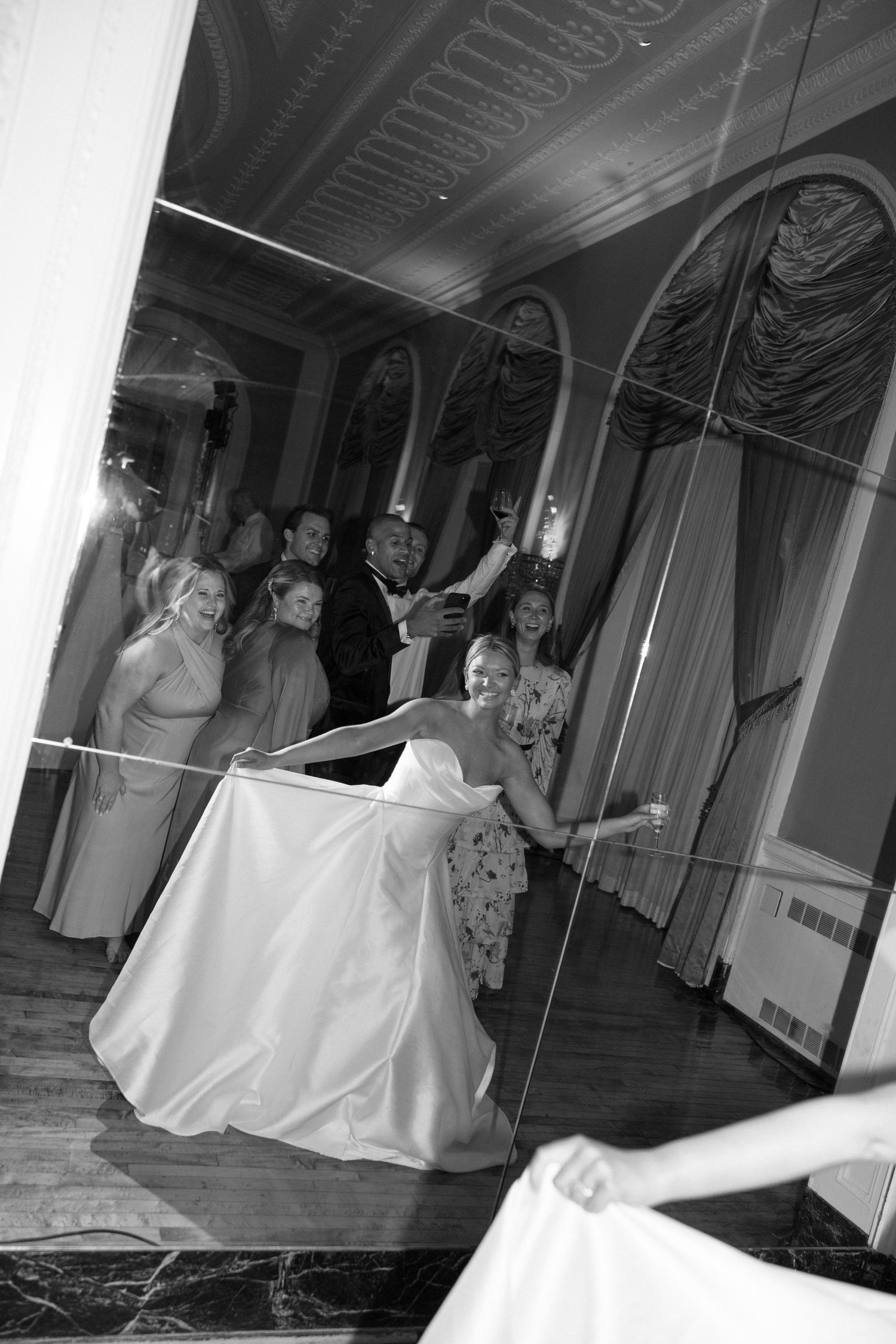 Black and white photo of a bride in a white gown dancing with guests at an elegant wedding reception venue.