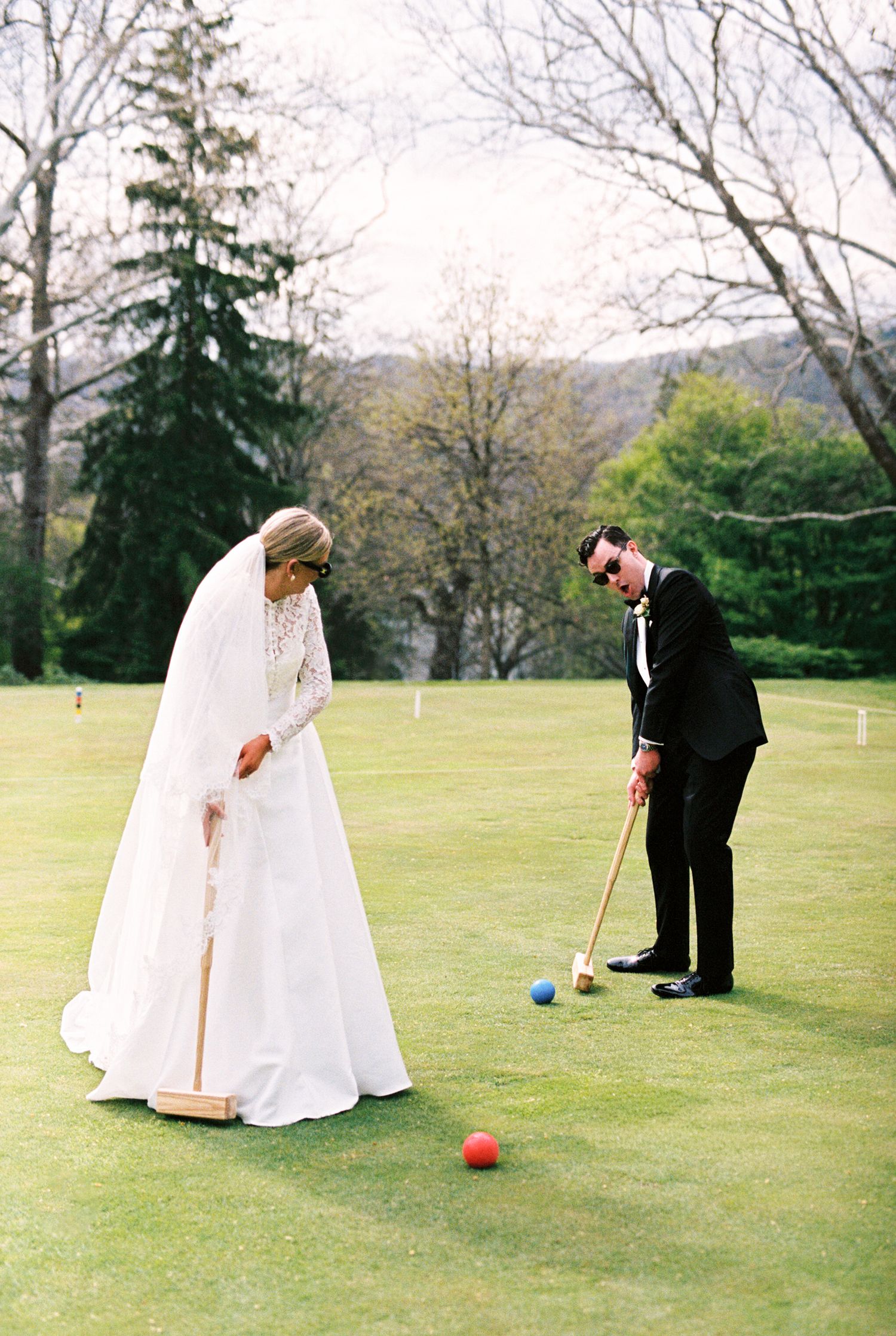 Bride and groom playing croquet on lush green lawn in wedding attire, with colorful balls on grass.