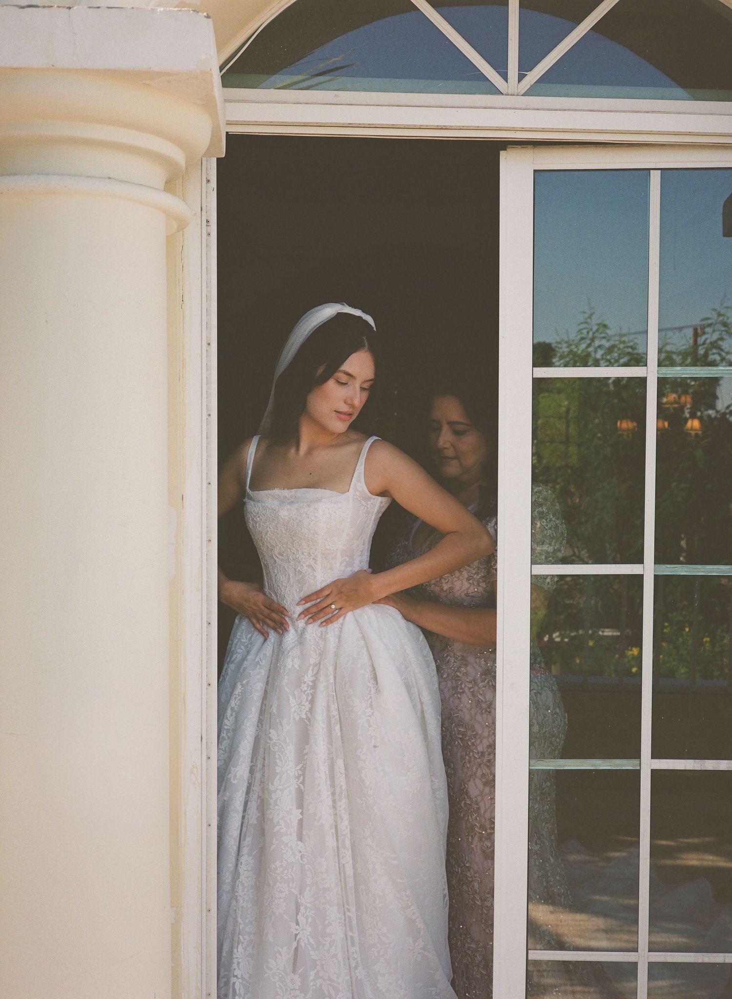 A woman in a white eyelet lace dress poses gracefully against white columns in natural sunlight.