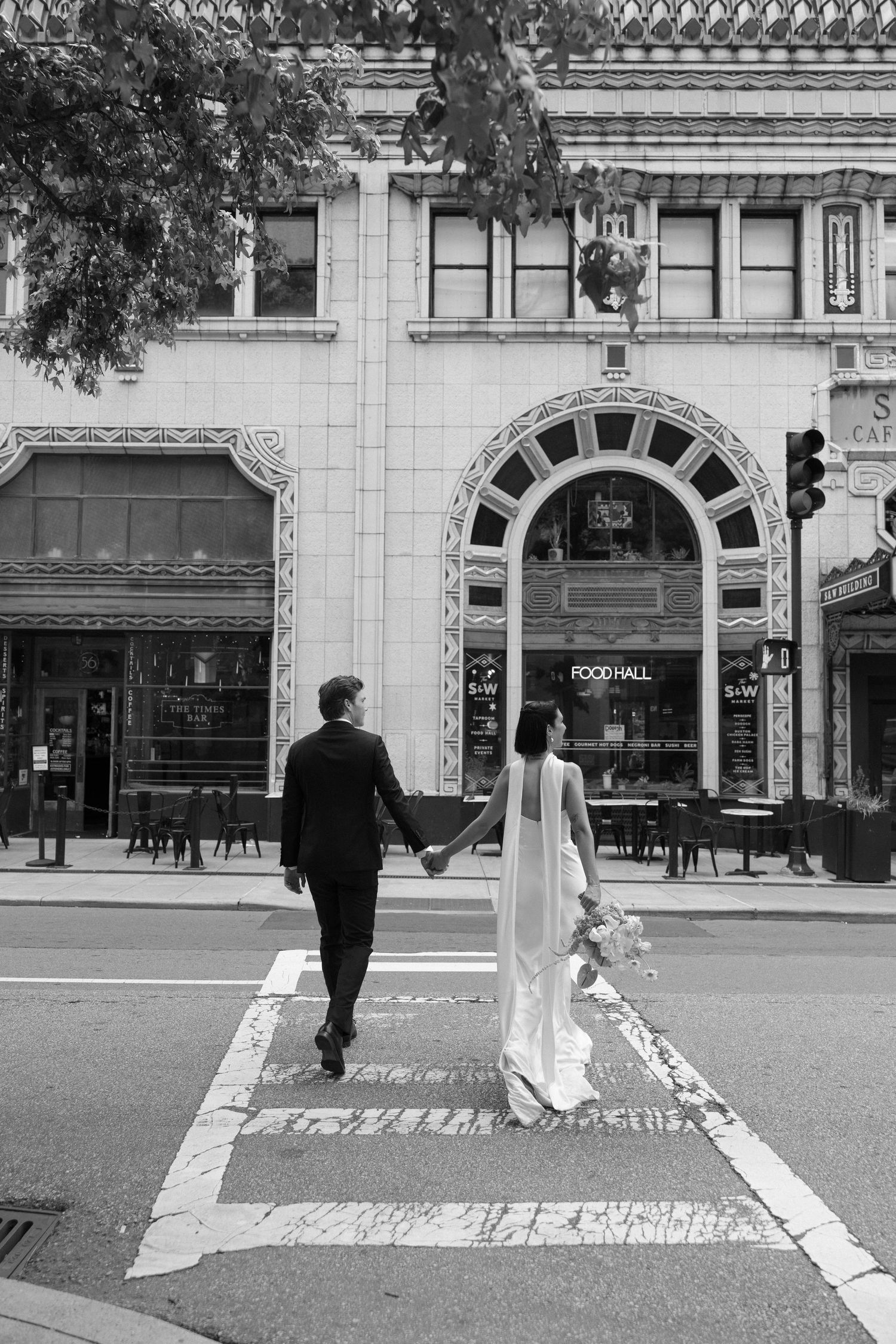 Bride and groom hold hands while crossing a city crosswalk toward an ornate Food Hall building in a black and white wedding photo.