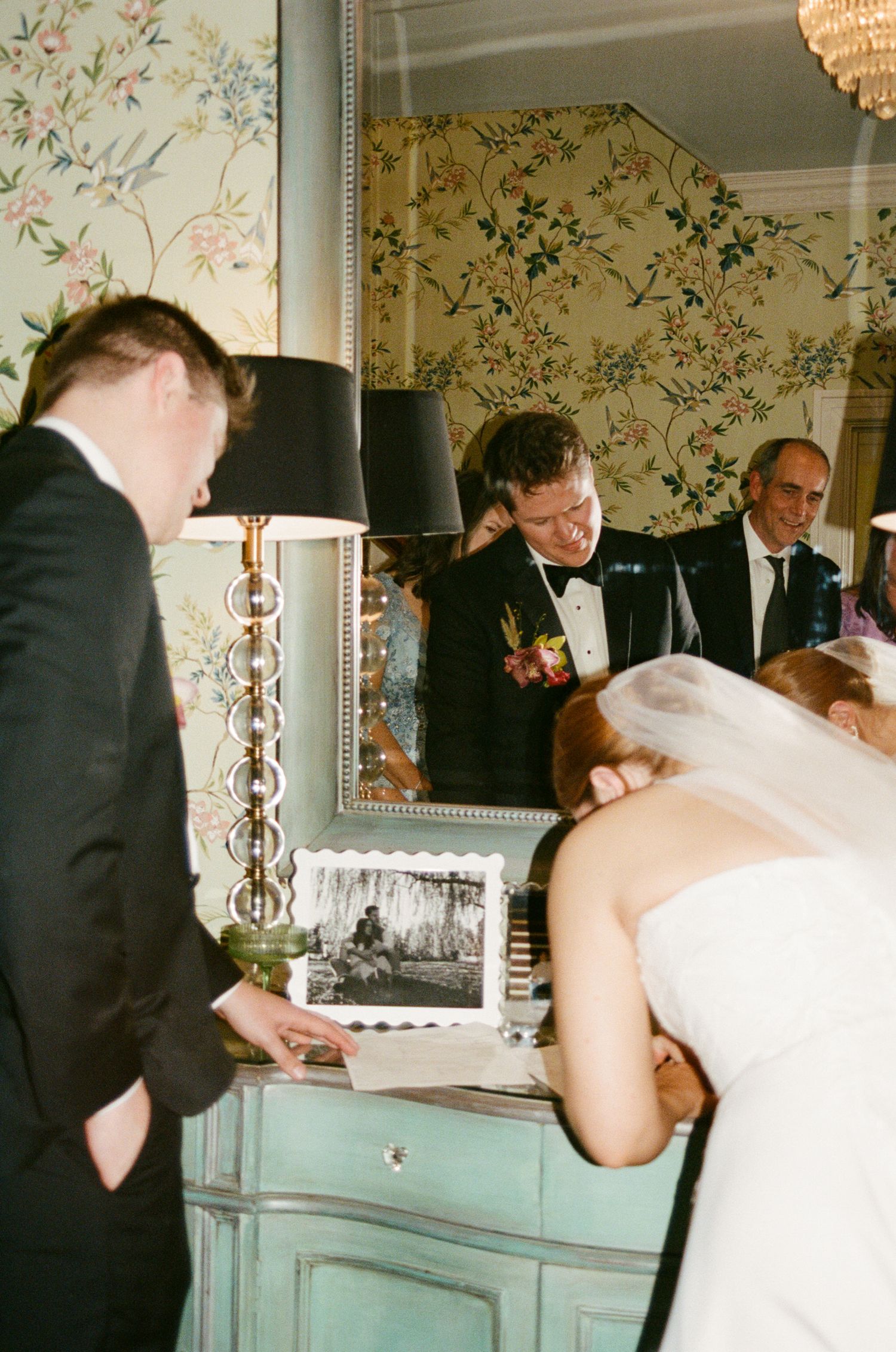 Bride and groom at vintage dresser with mirror reflection showing wedding guests in floral wallpapered room.