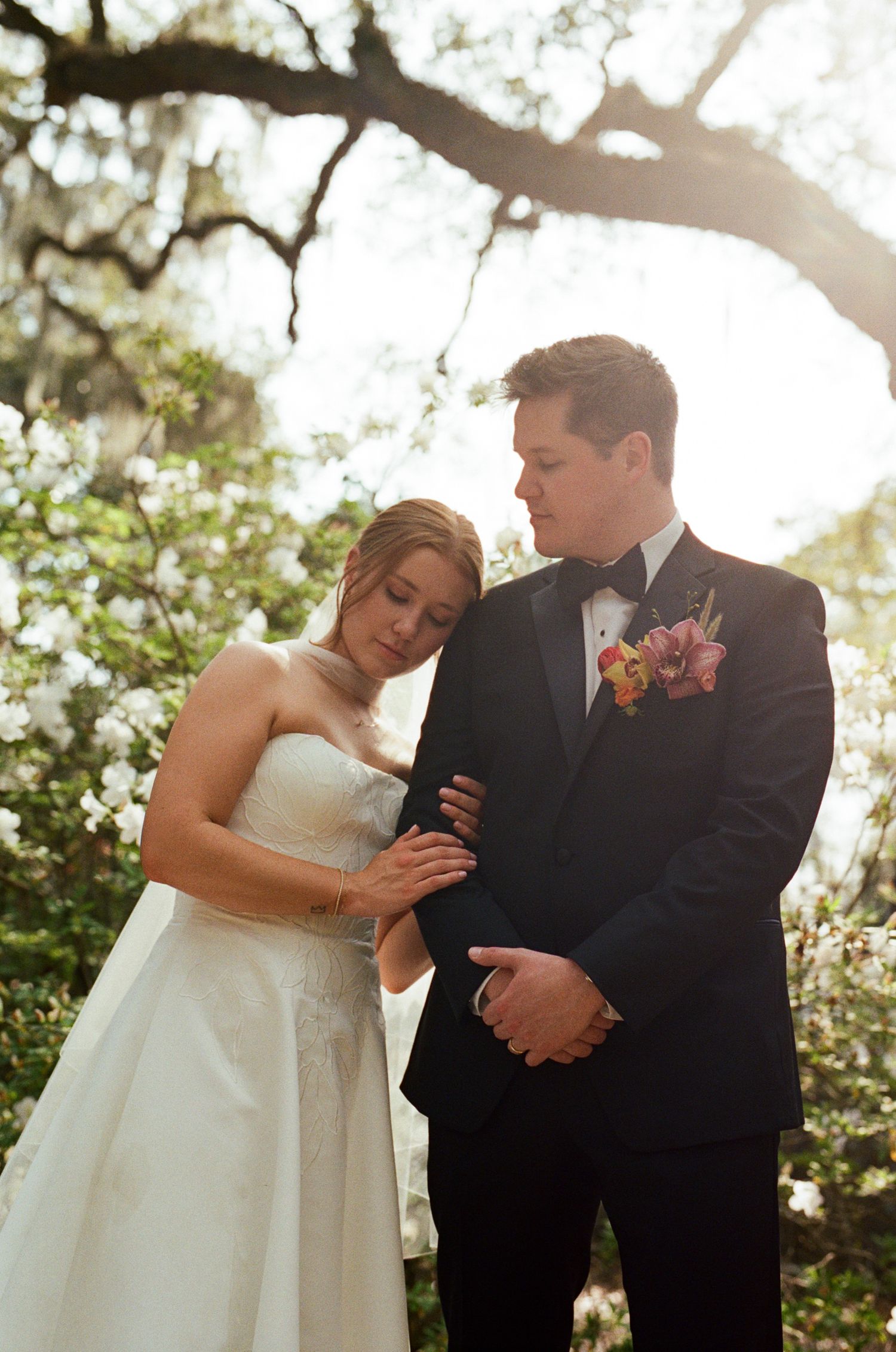 Bride and groom share an intimate moment outdoors, she in a strapless gown, he in a black tuxedo with floral boutonniere.