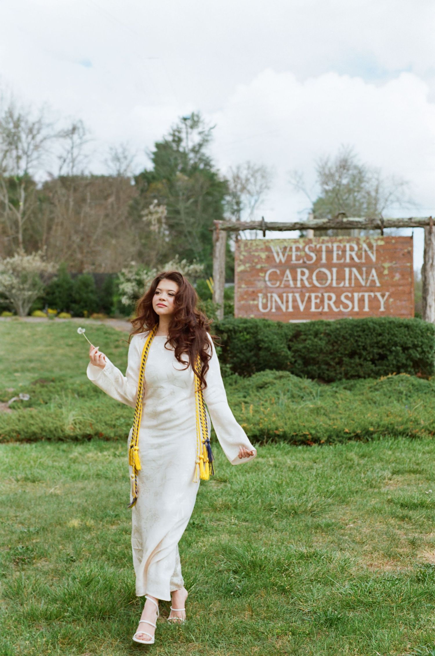 A young woman in a white dress with yellow graduation sash poses in front of Western Carolina University sign.