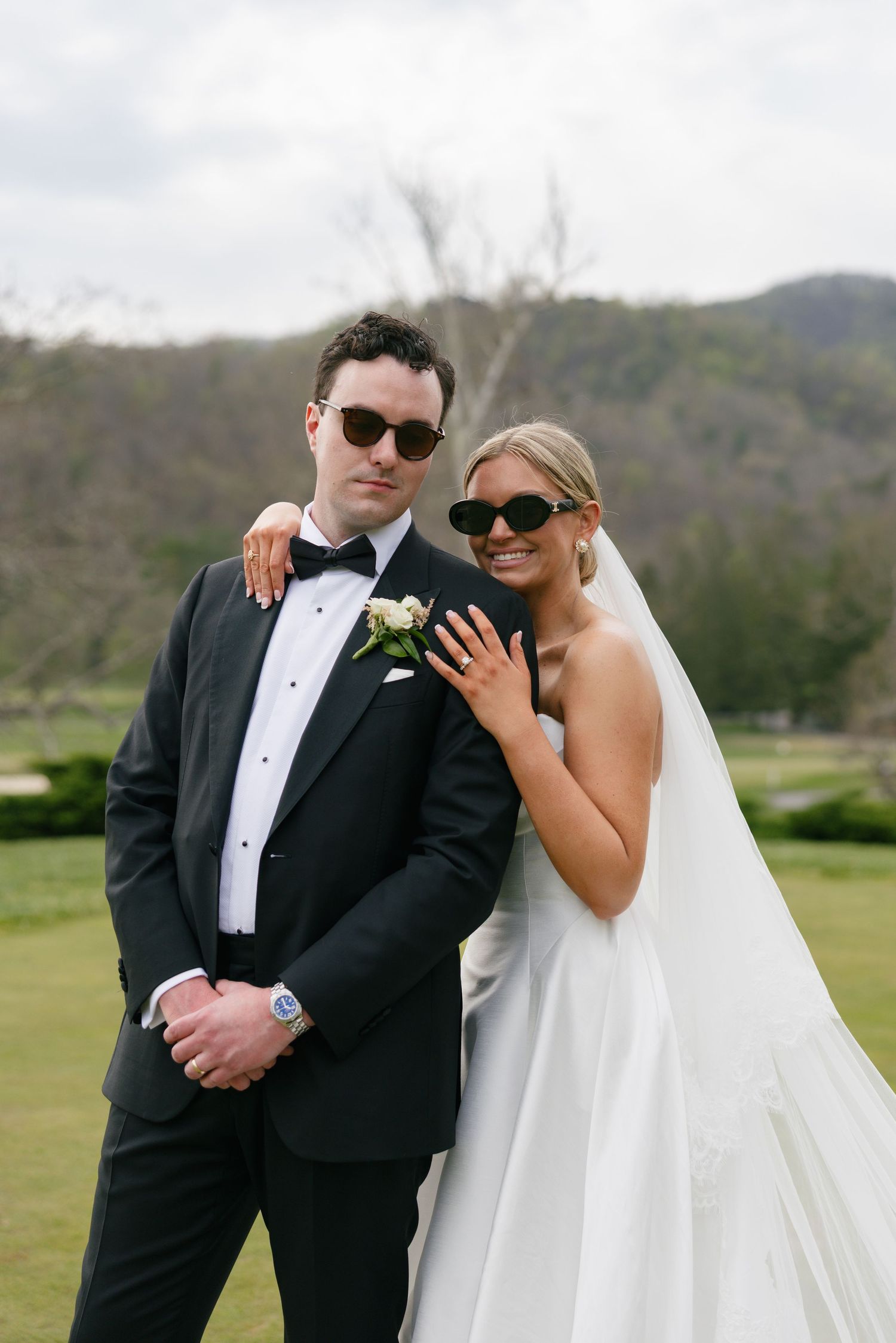 A bride and groom pose outdoors in wedding attire and sunglasses, with green hills in the background.