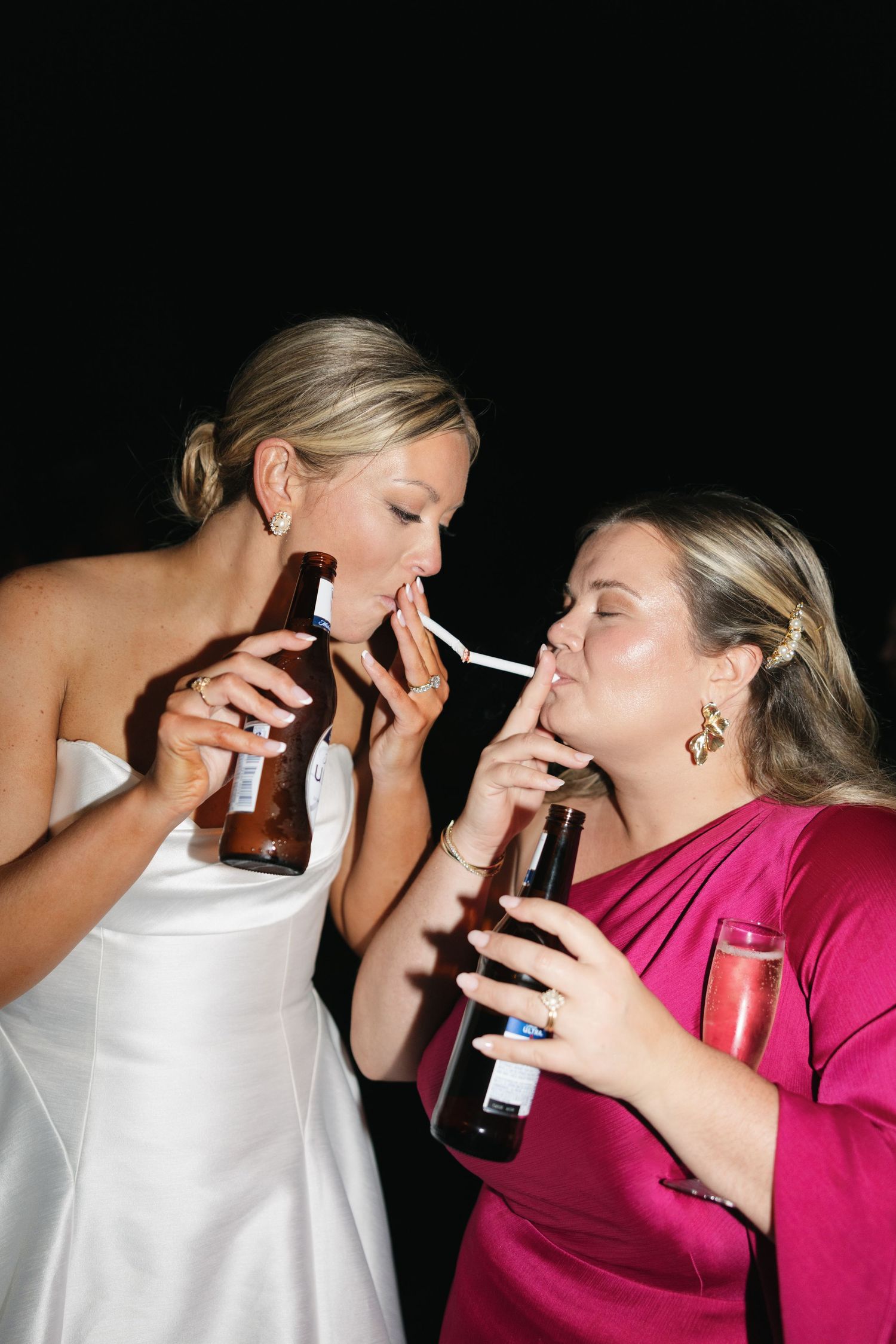 Two women in formal attire sharing a cigarette light while holding beer bottles at a wedding celebration.