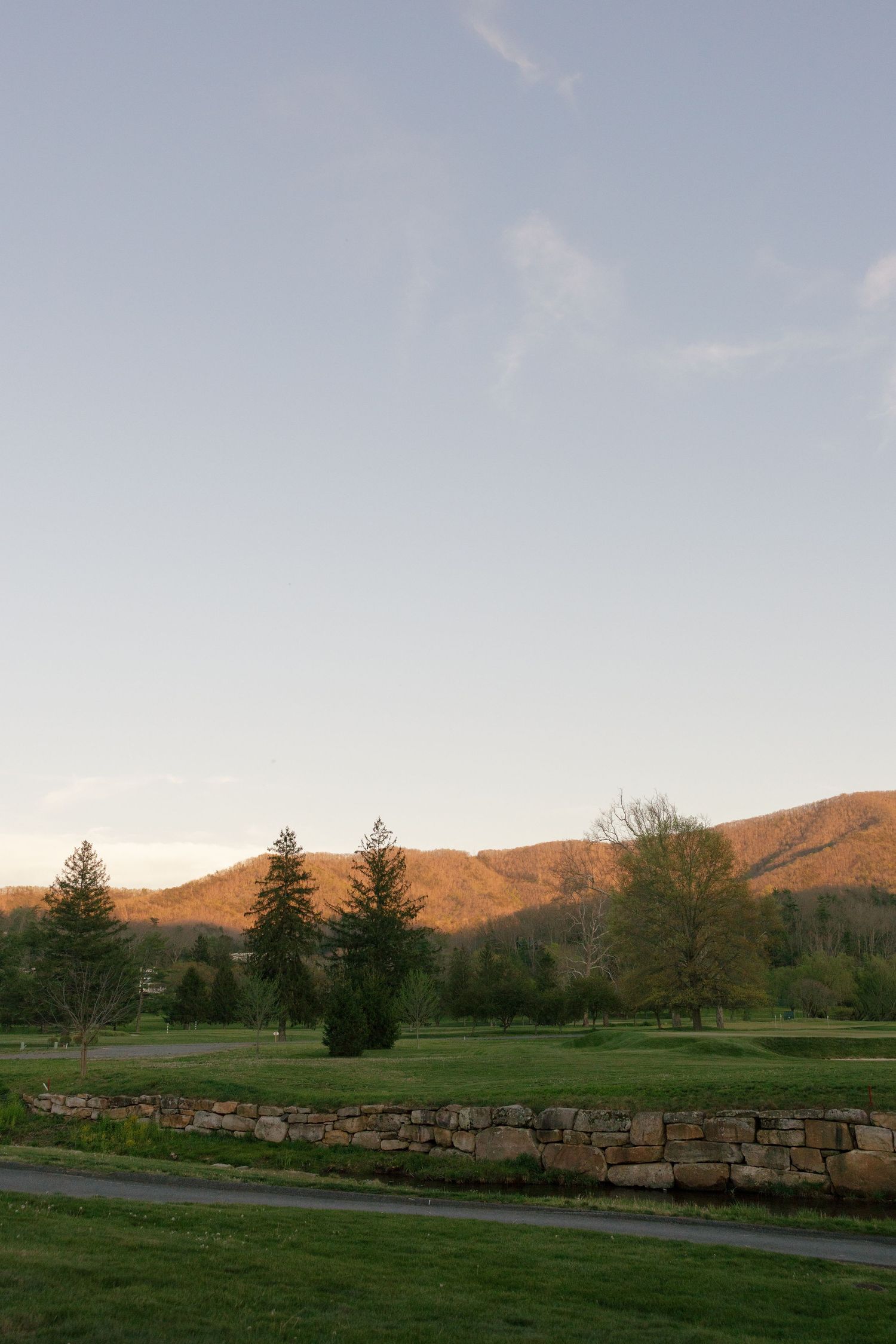 A serene golf course landscape at sunset with green grass, stone wall, evergreen trees, and golden mountains under a clear sky.