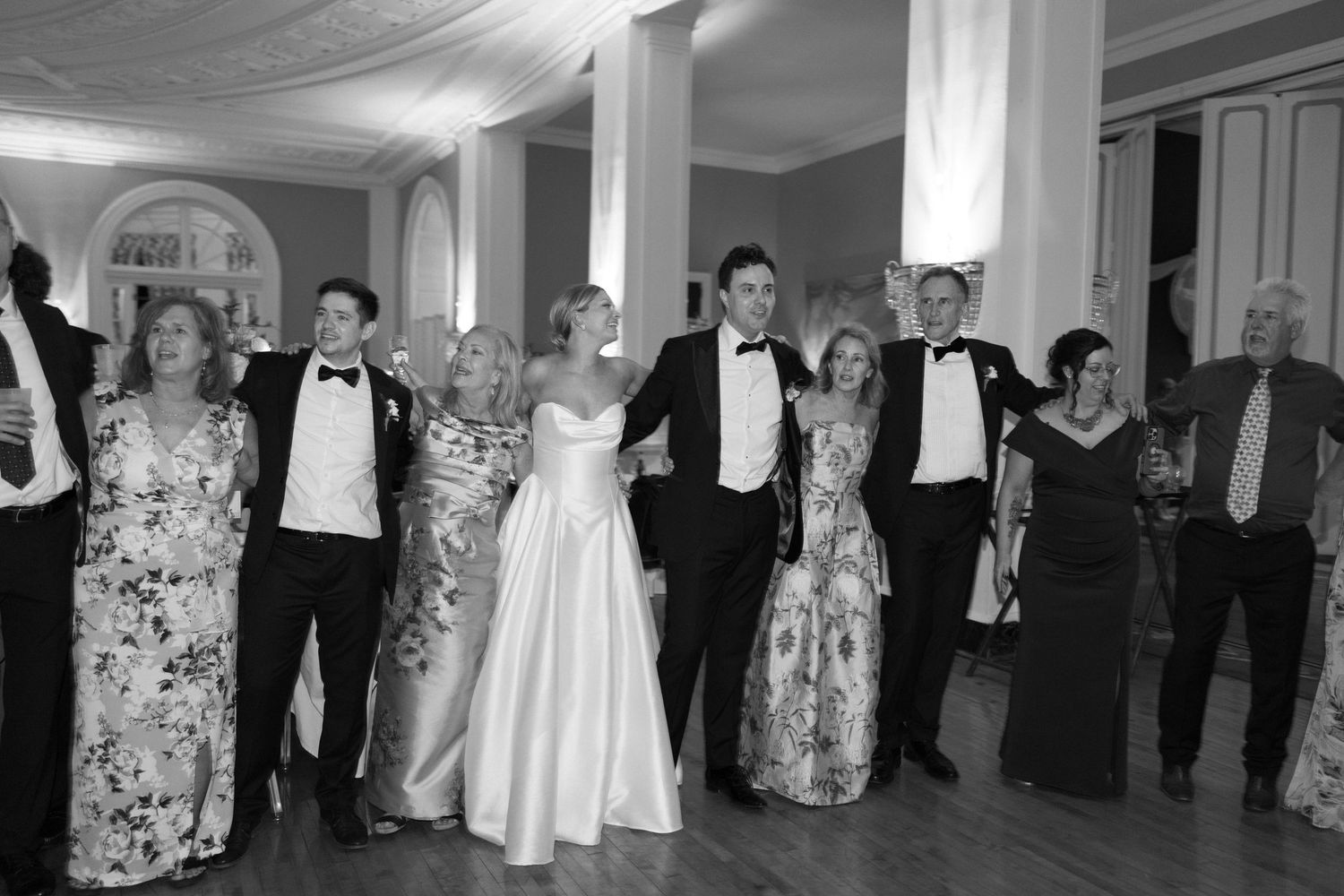 Black and white photo of a wedding party in formal attire smiling together on a dance floor.