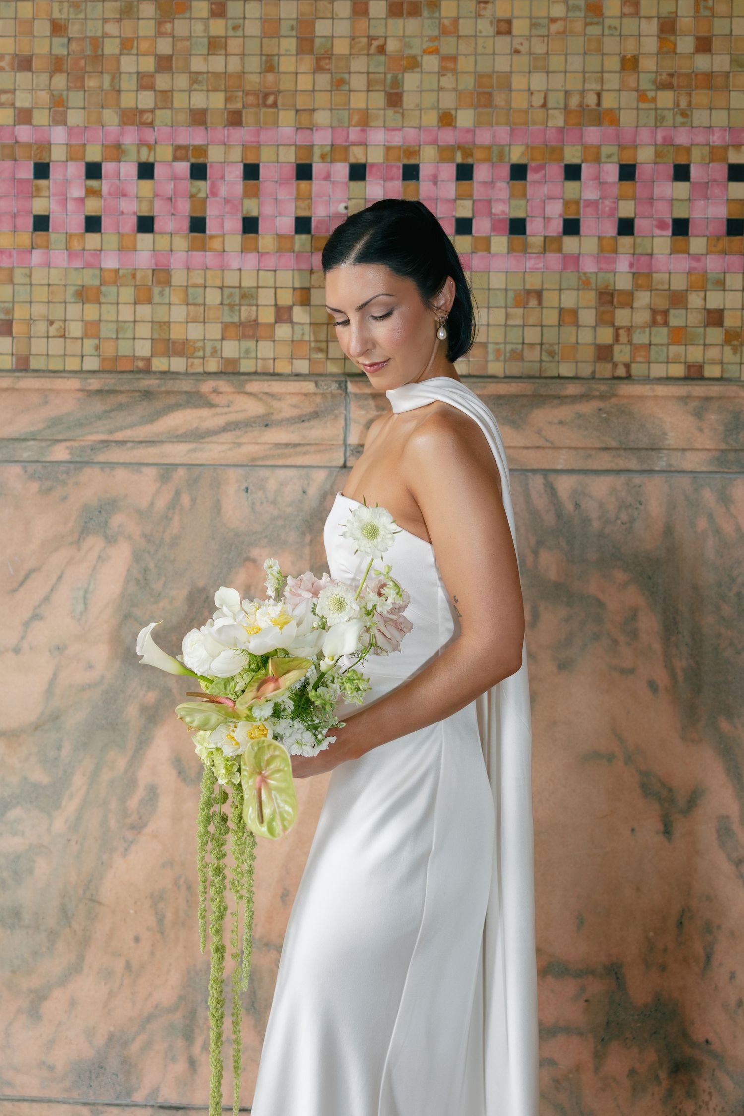 Bride in an elegant white satin gown holding a cascading green and white bouquet in front of pink marble and mosaic tile wall.