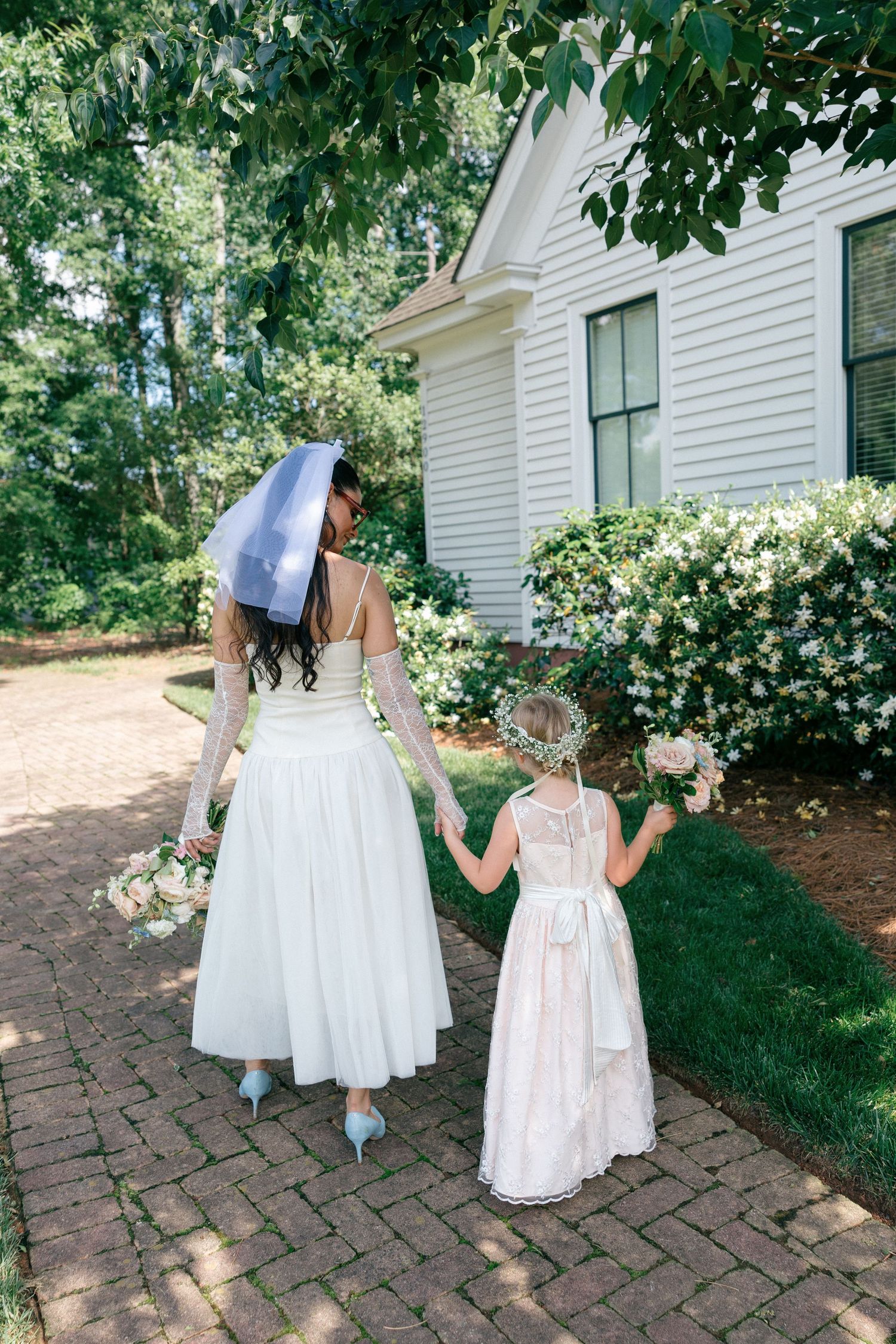 Bride in a white tea-length dress and short veil walks hand in hand with a flower girl holding a bouquet along a brick path.