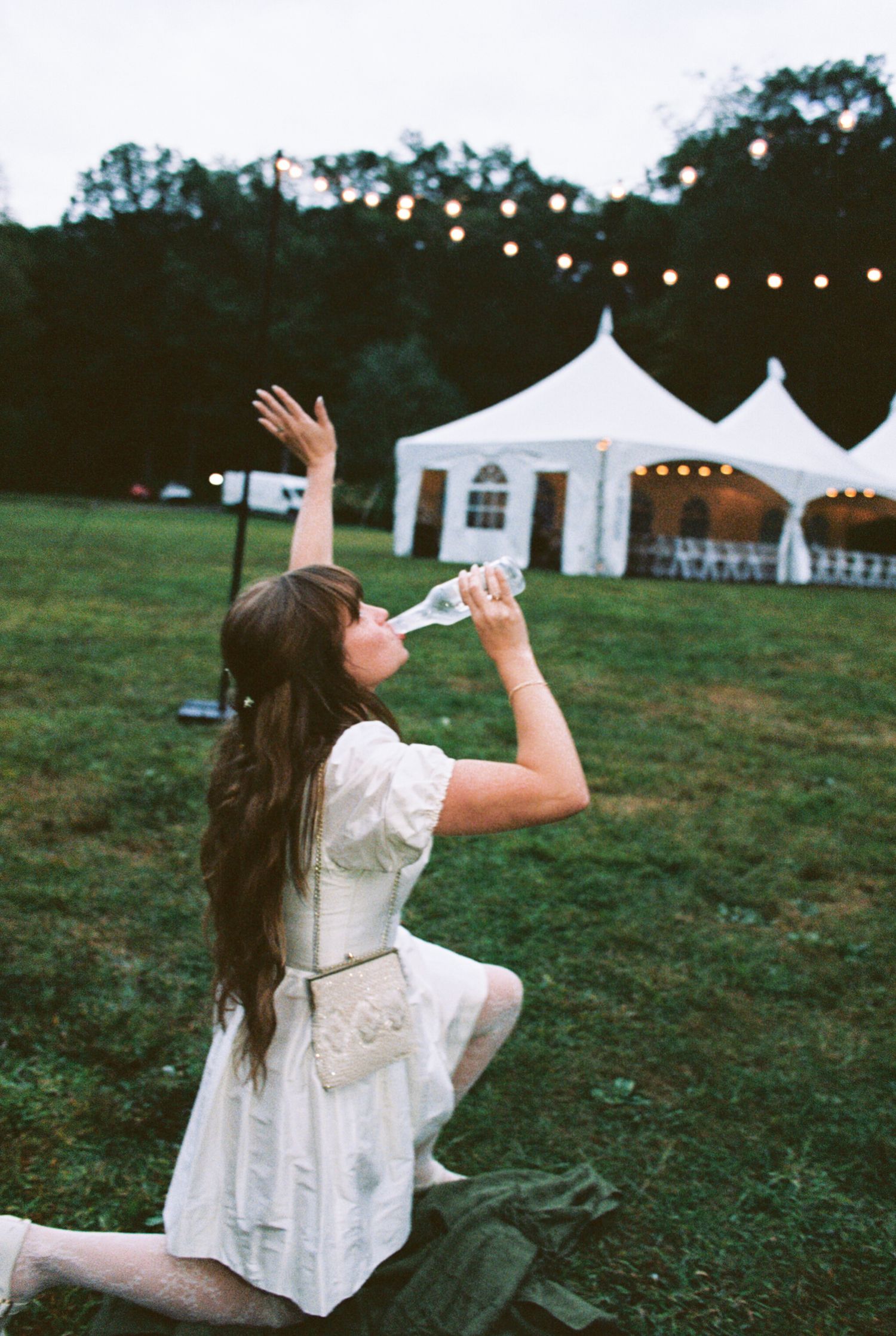 A figure in a flowing white dress dances in a field at dusk with string lights and white tents in the background.