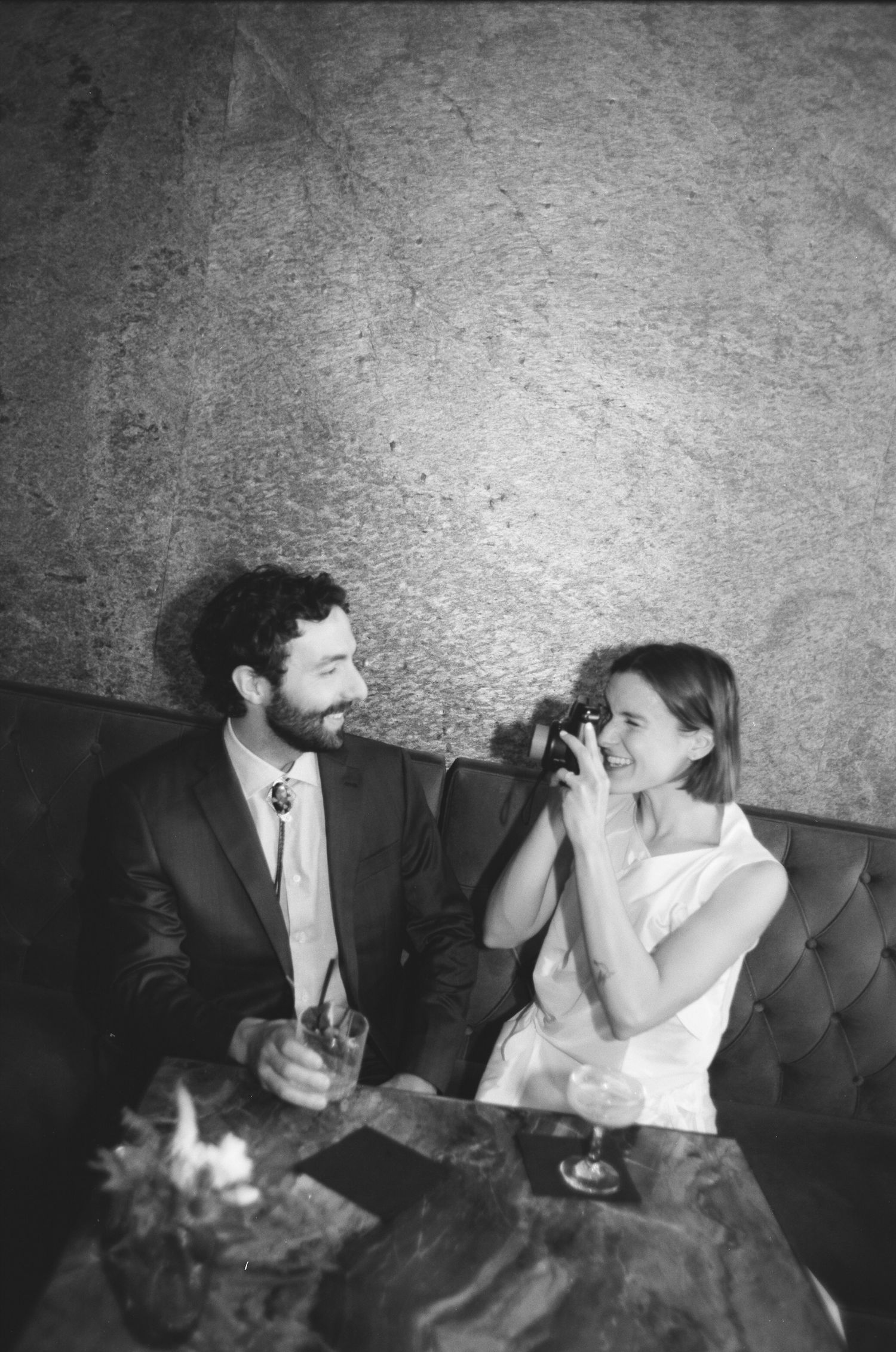 Black and white photo of a smiling couple sitting at a table, man in suit holding drink, woman with camera at wedding.
