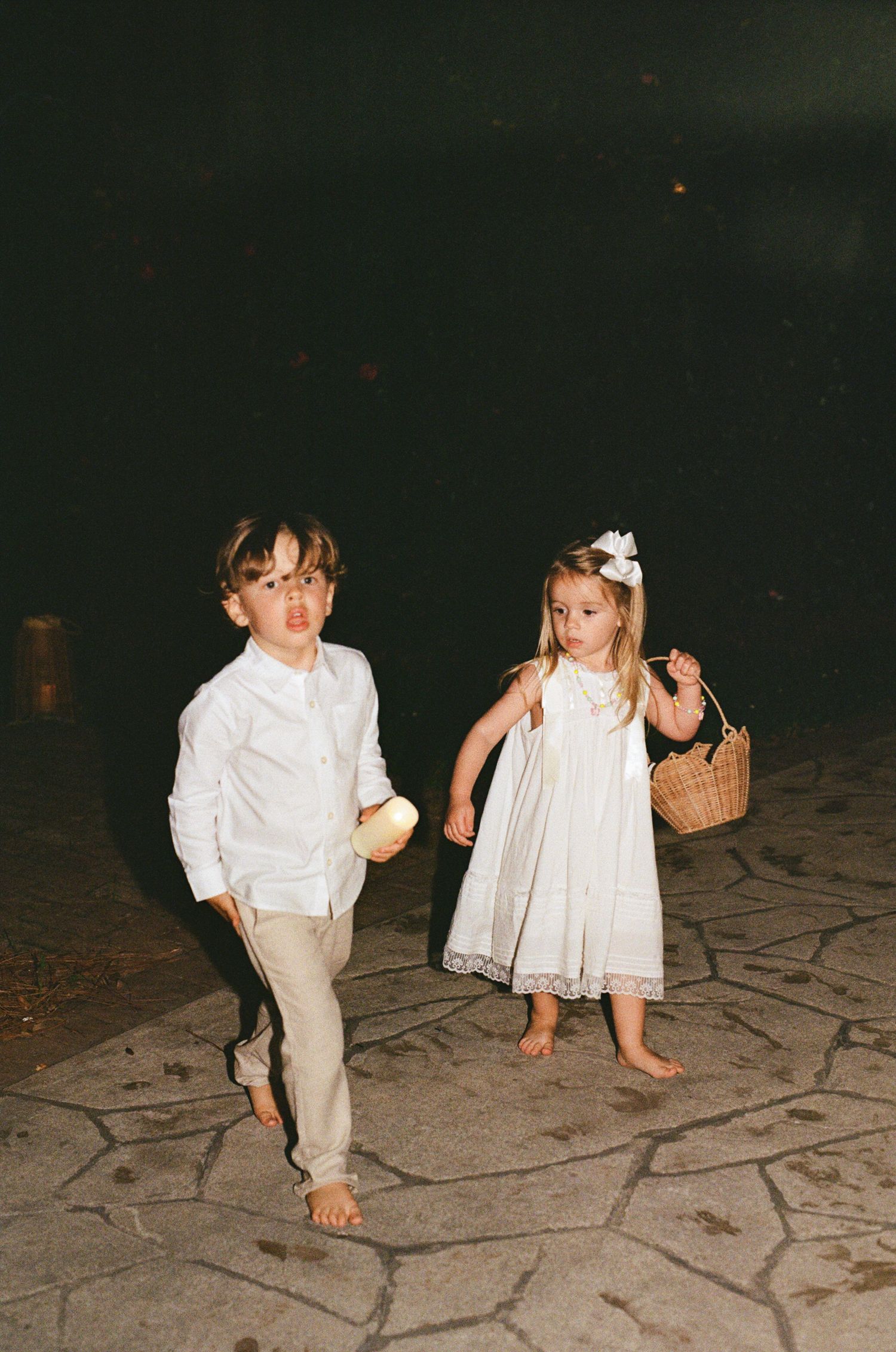 Two barefoot children in white wedding attire walk at night, a boy in shirt and pants and a girl in dress holding a basket.
