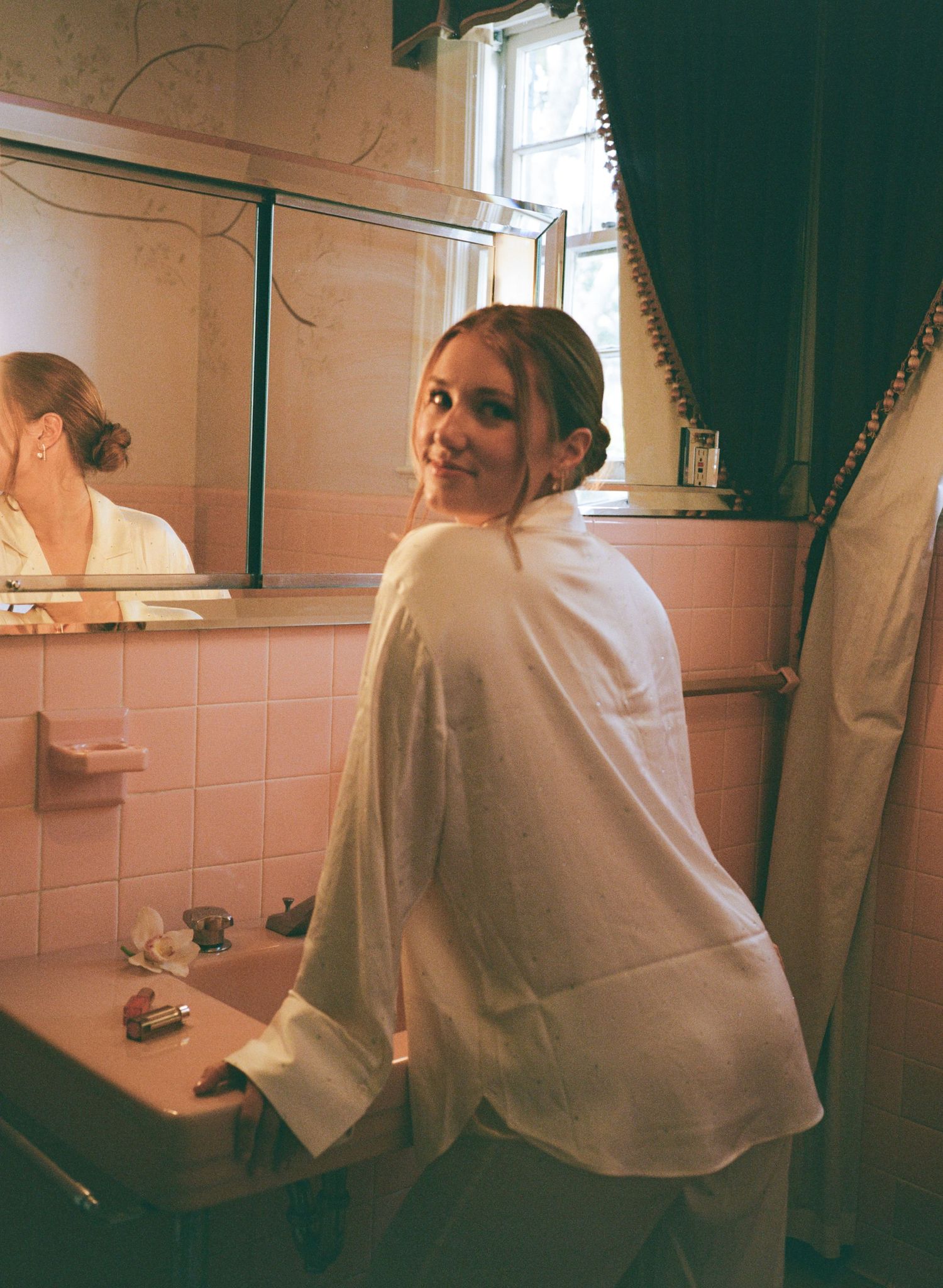Woman in white robe smiling over shoulder in vintage pink tiled bathroom with mirror reflection.