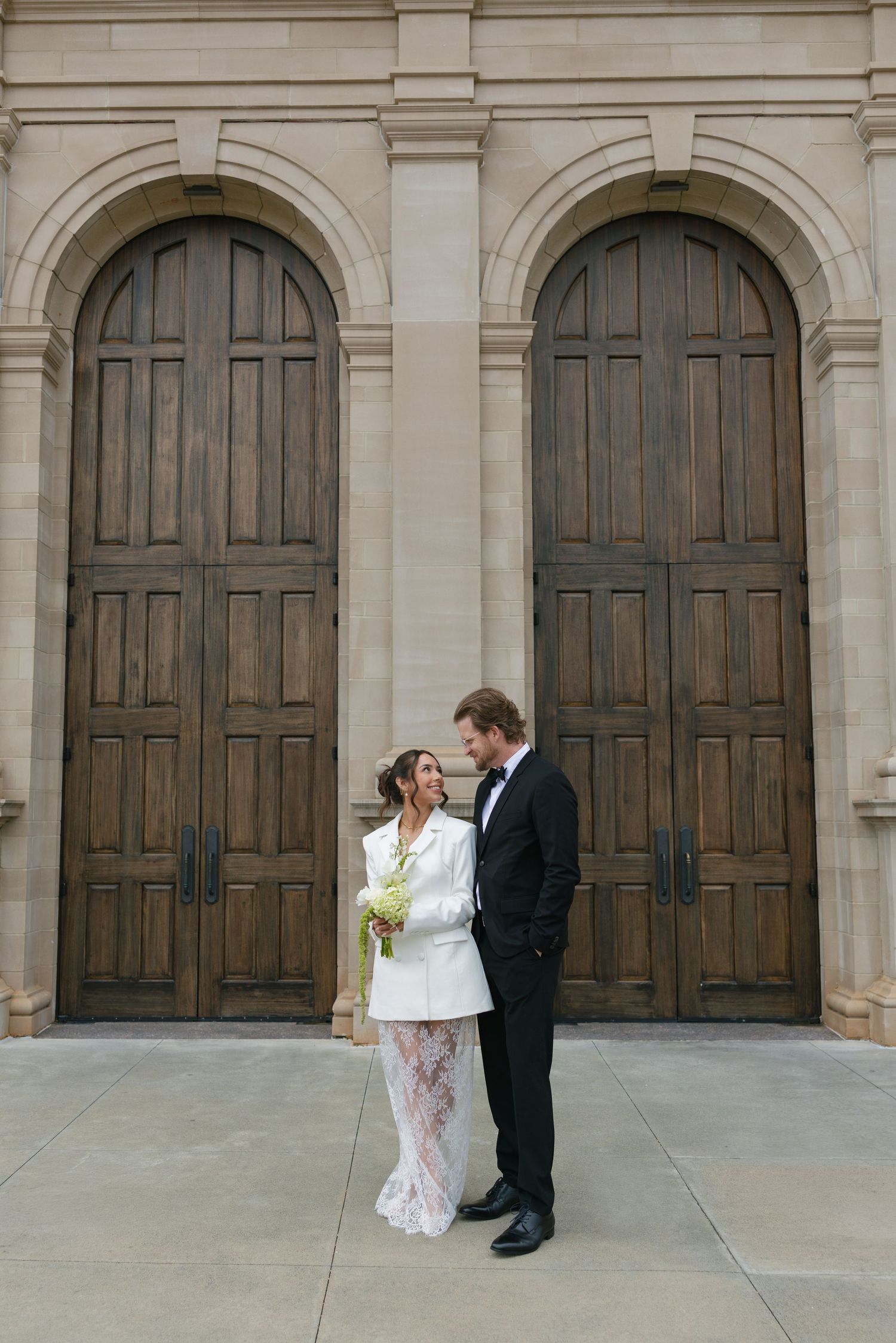 Bride in white blazer and lace skirt holds bouquet beside groom in black tuxedo before arched wooden doors.