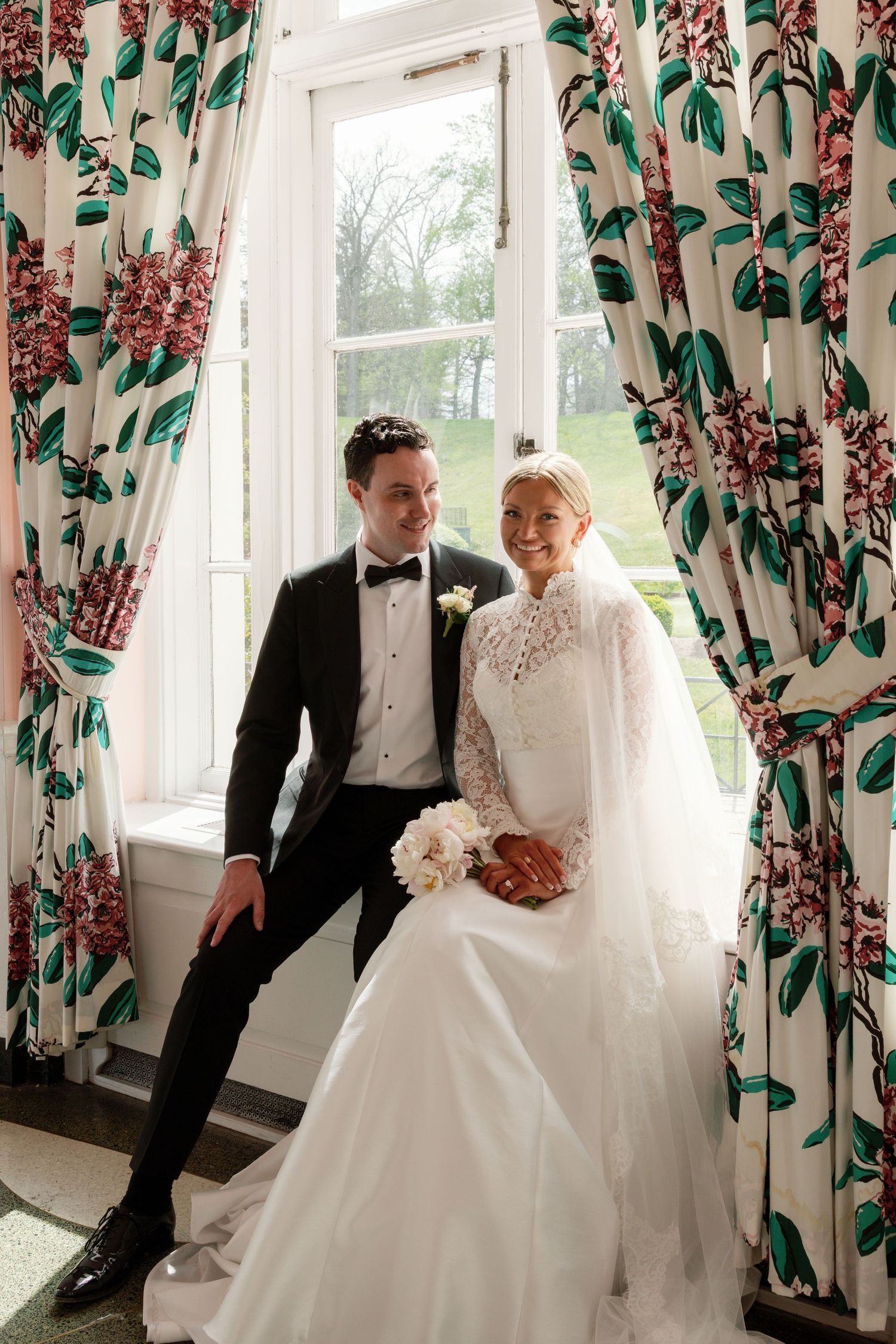 Bride in white lace gown and groom in black tuxedo pose by floral curtained window on their wedding day.