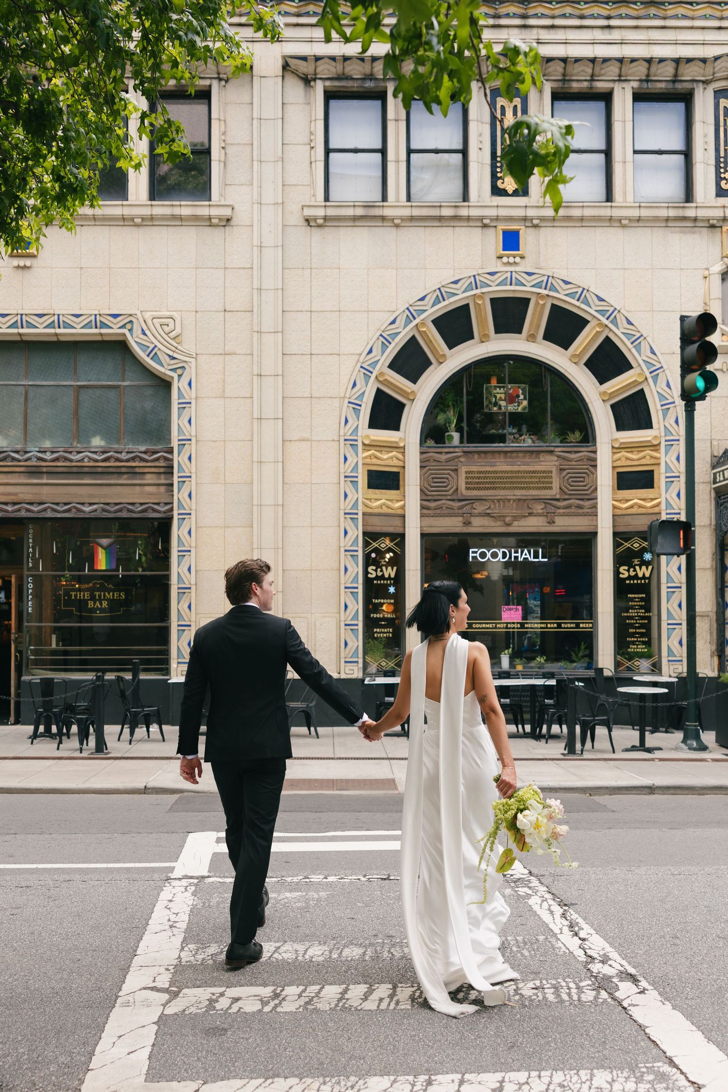 Bride in a flowing white gown and groom in a black suit hold hands while crossing a city street in front of an ornate Food Hall entrance.
