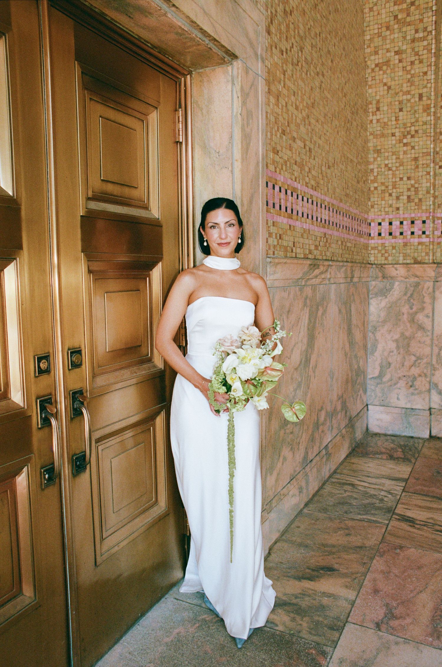 Bride in a strapless white gown and choker holding a pastel bouquet beside ornate bronze doors in a pink marble hallway.