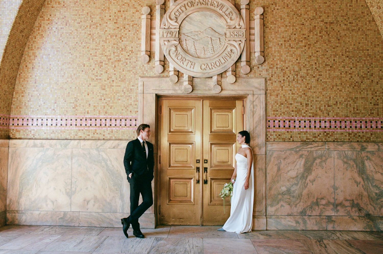 Bride in a white gown holding a bouquet and groom in a dark suit stand beside ornate golden doors in a marble-walled historic wedding venue.