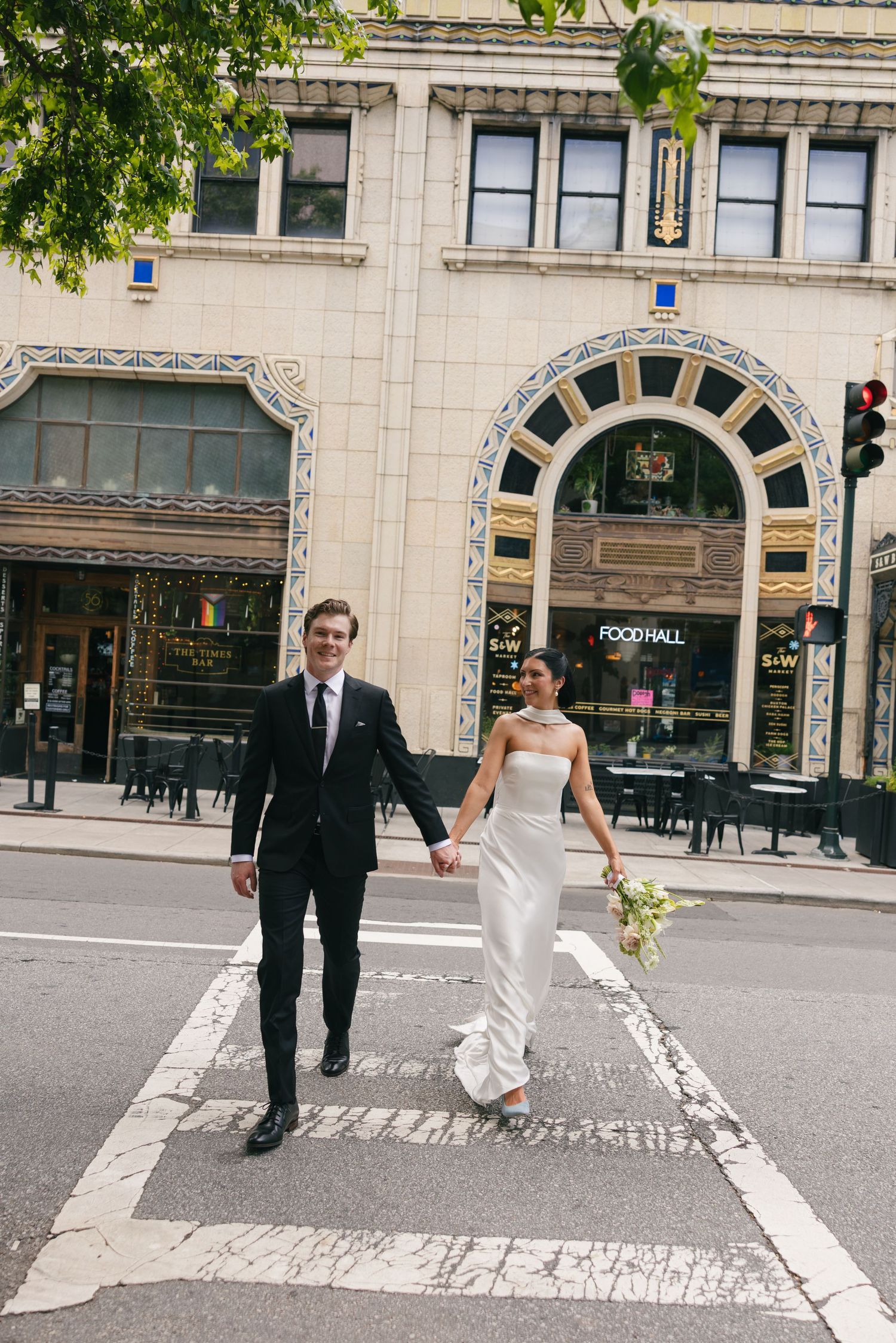 Bride in a white strapless gown and groom in a black suit hold hands while crossing a city street past an ornate vintage building.