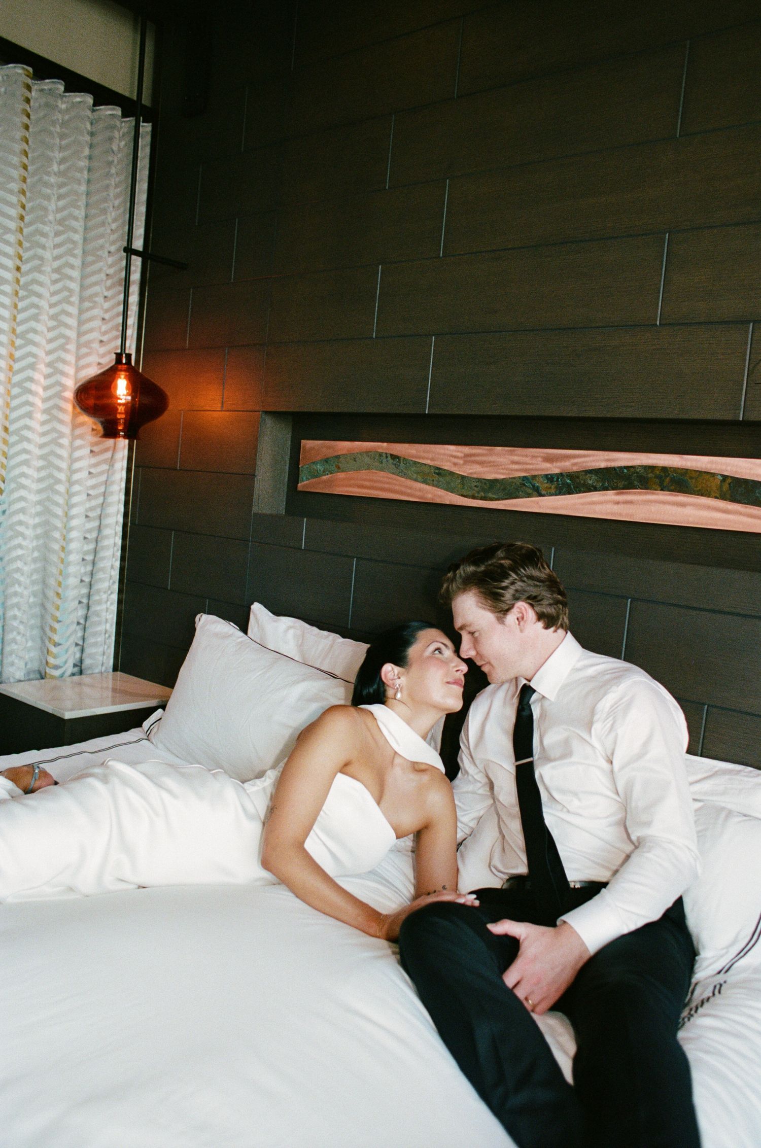 Bride in a white gown and groom in a white shirt and black tie share an intimate moment on a bed in a modern hotel room.