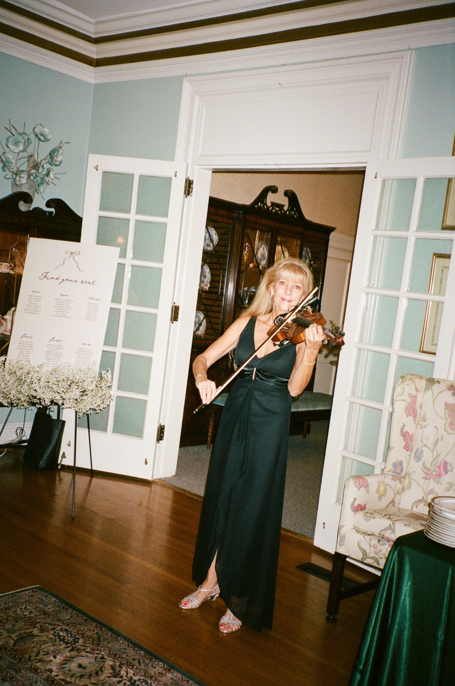 Woman in a long green dress playing the violin in an elegant room with French doors, a seating chart, and floral armchair.